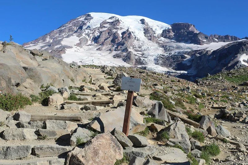 Trail with rocks and wooden steps leading towards a snow-capped mountain, with a sign that reads "Allez Vista" in the foreground.