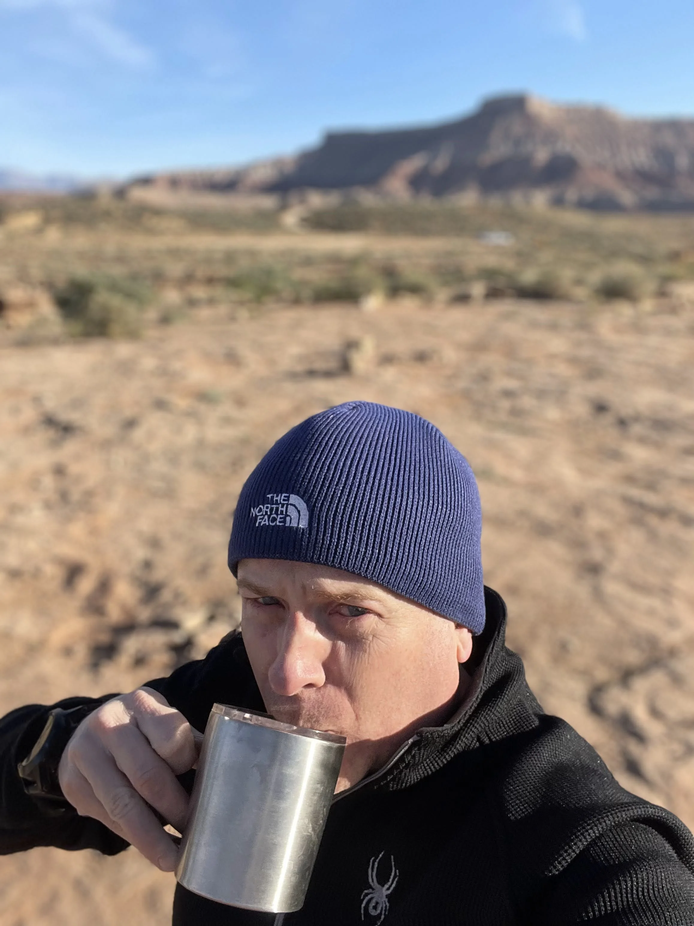 A person wearing a blue The North Face beanie and black jacket holds and drinks from a metal mug in a desert landscape with rocky formations in the background.