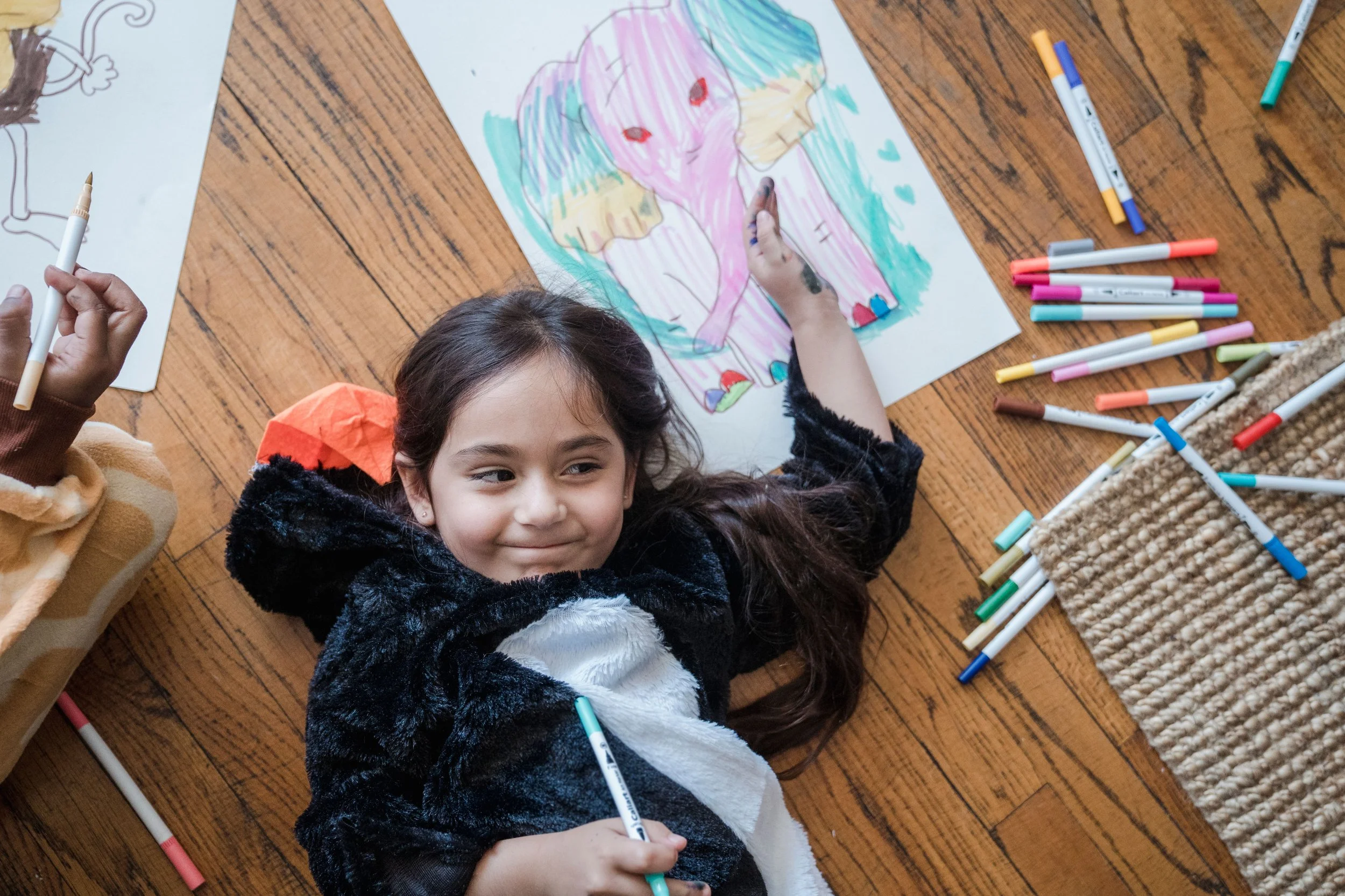 An elementary school aged child with light skin and long brown hair laying on the floor surrounded by drawings and markers