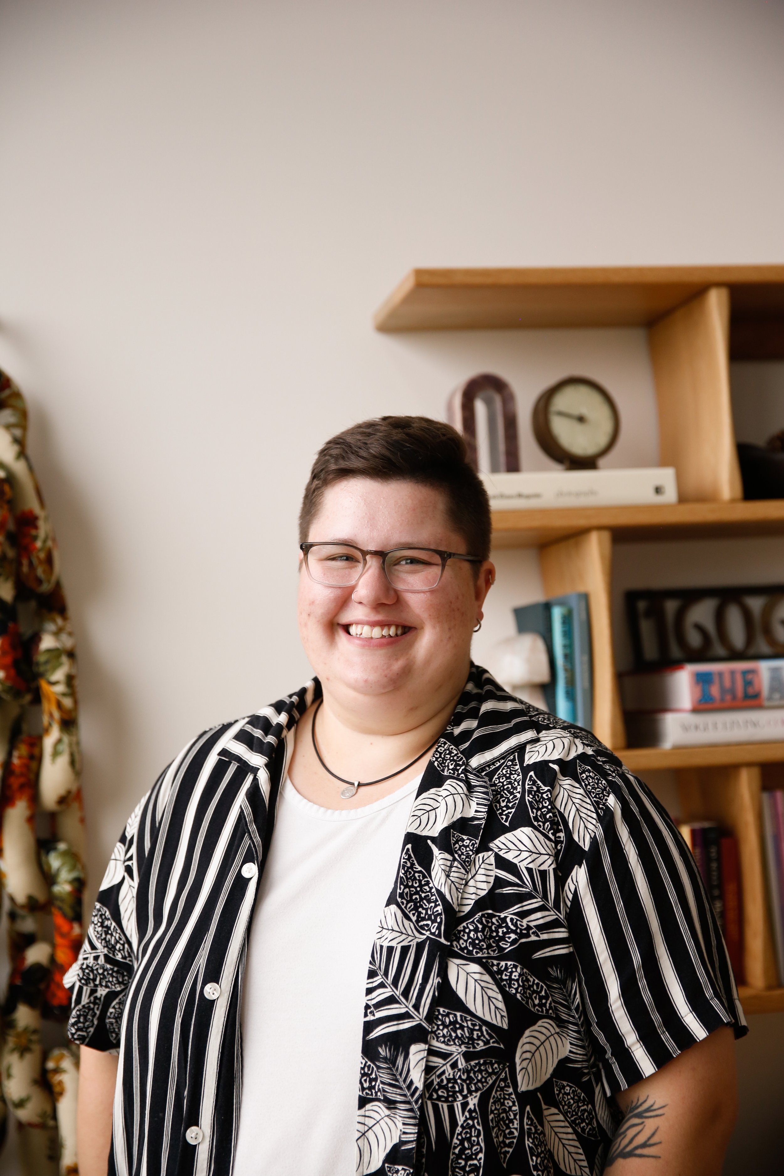 Laura Kent is pictured here smiling. They are a fat white person with short straight brown hair and glasses. They are wearing a black and white striped shirt with leaves on it.