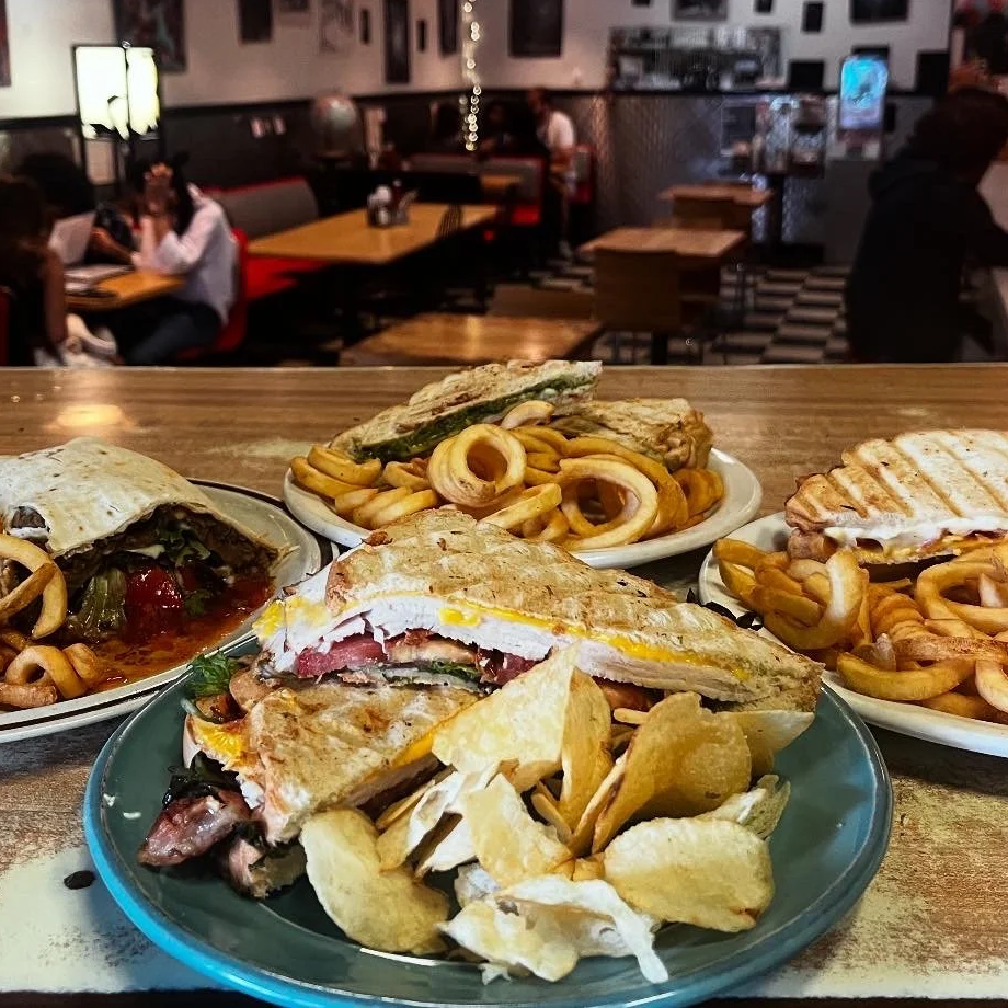 A variety of sandwiches, salads, and potato chips on plates on a wooden table in a restaurant with people dining in the background.