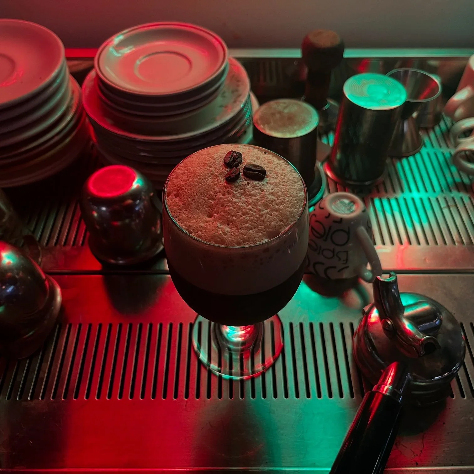 A frothy coffee drink with coffee beans on top in a wine glass, surrounded by coffee cups and plates on a metallic tea or coffee station with colored lighting.