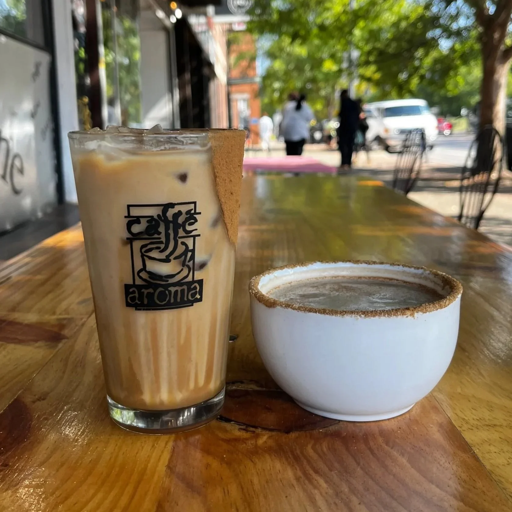 A glass of iced coffee with a cookie on the side and a white coffee cup on a wooden table outdoors, with blurred people and trees in the background.