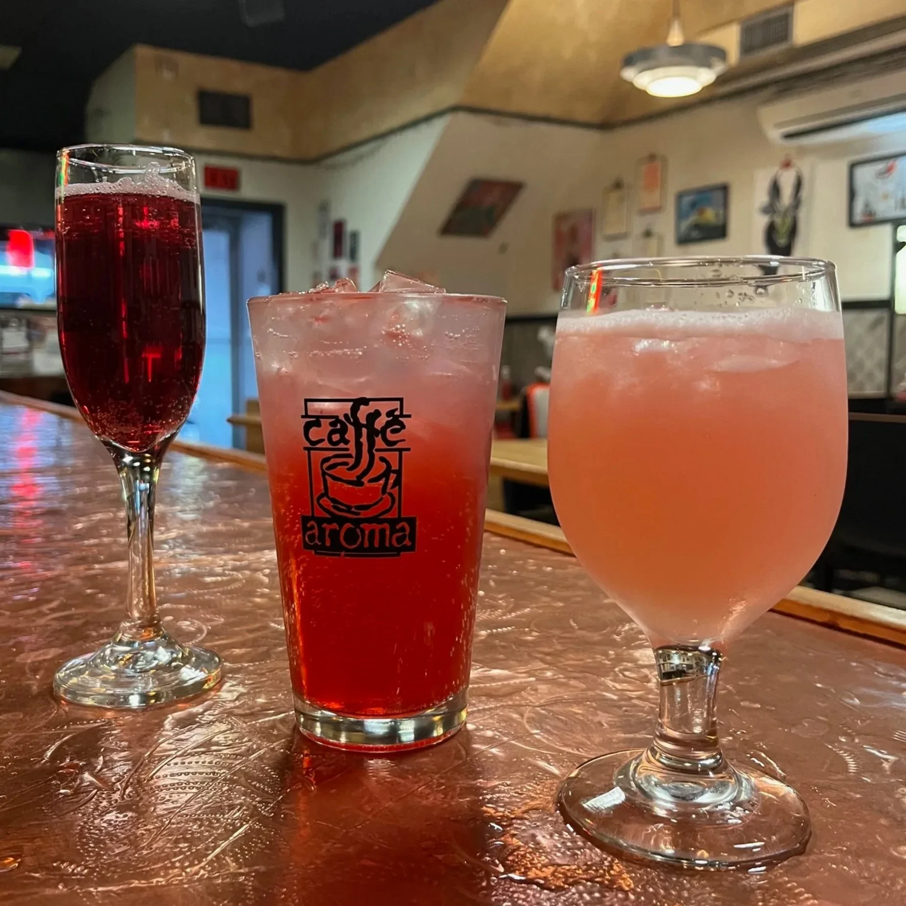 Three glasses of drinks on a table inside a restaurant. The left glass is a tall champagne flute filled with red beverage. The middle glass is a large tumbler with a pinkish drink and ice. The right glass is a large stemmed wine glass with a pinkish drink and ice. The background shows a wall with framed pictures and decorative artwork.