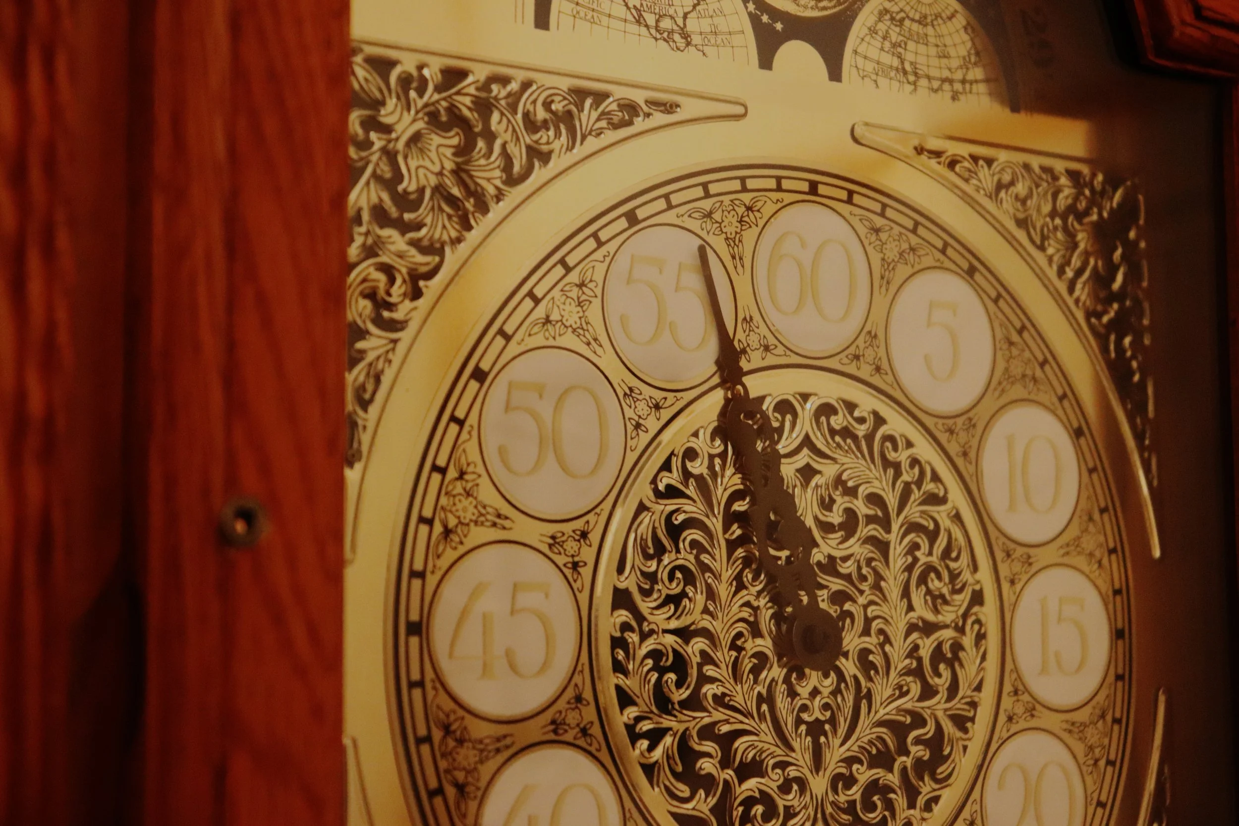 Close-up of a vintage clock face with black ornate designs and numbers, partially obscured by a wooden frame.