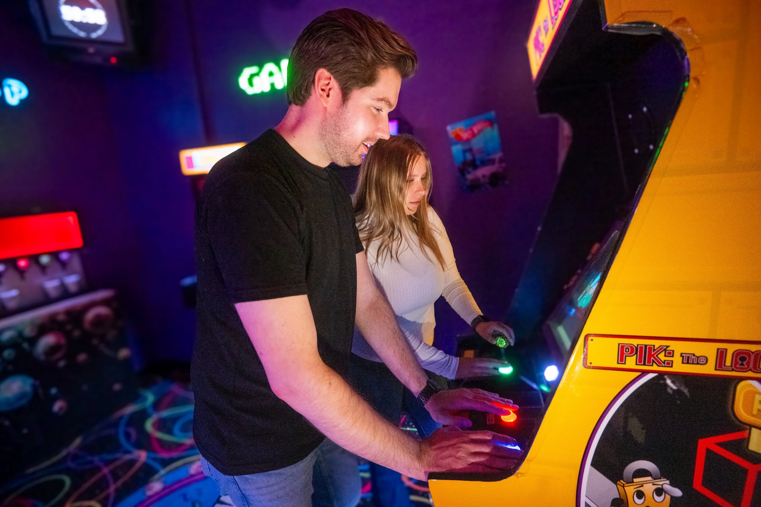 Two people playing an arcade game with a yellow Arcade machine, in a room with neon lights and colorful carpet.