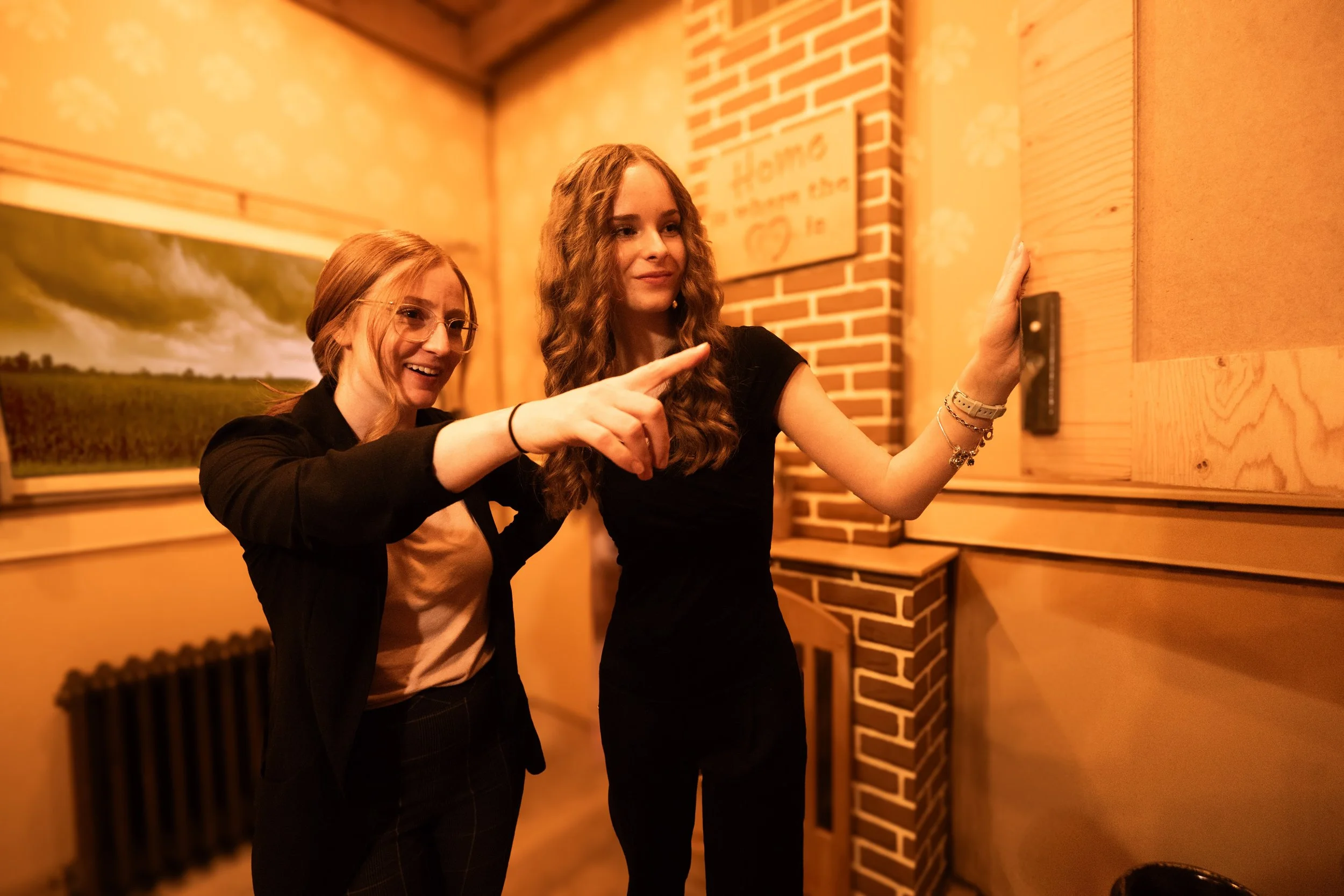 Two young women with curly and straight hair, one with glasses, pointing and looking at a wooden box on the wall in a warmly lit room with a brick and wood interior.