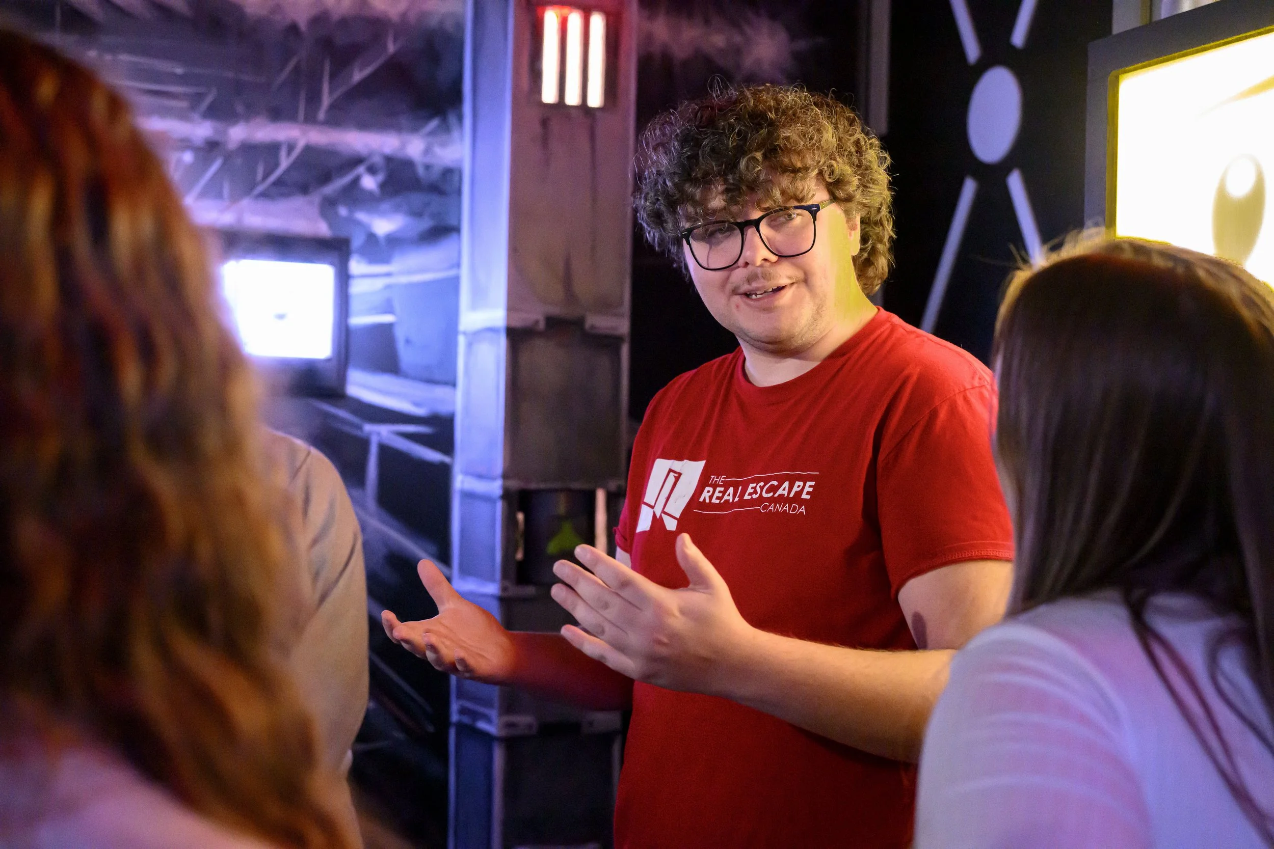 A man with curly hair and glasses wearing a red T-shirt with white text is speaking to a group of people in a dimly lit room with computer equipment and mounted screens.