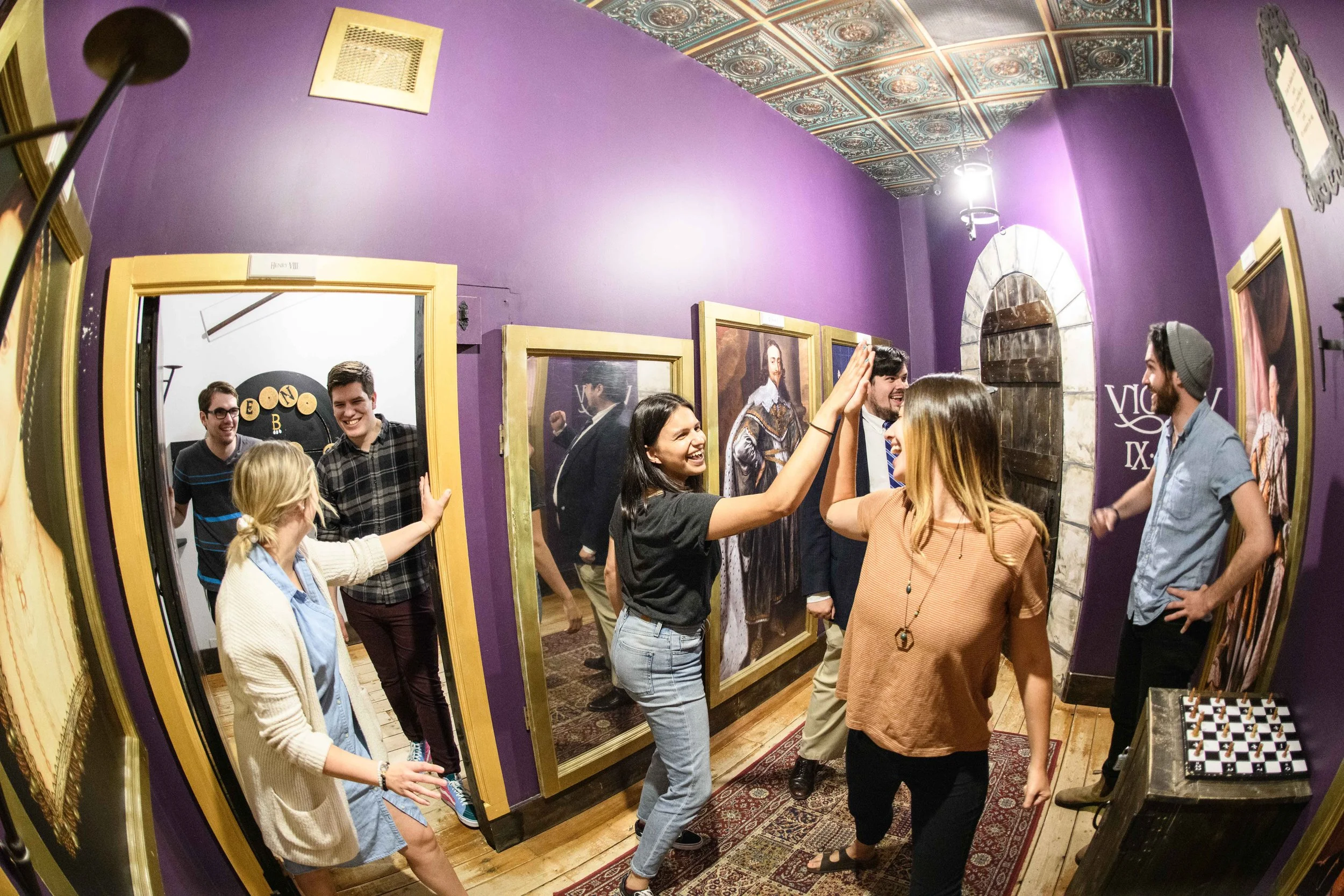 Group of young adults smiling and chatting in a cozy room with purple walls, decorated with framed portraits and vintage decor, through a large fish-eye mirror.