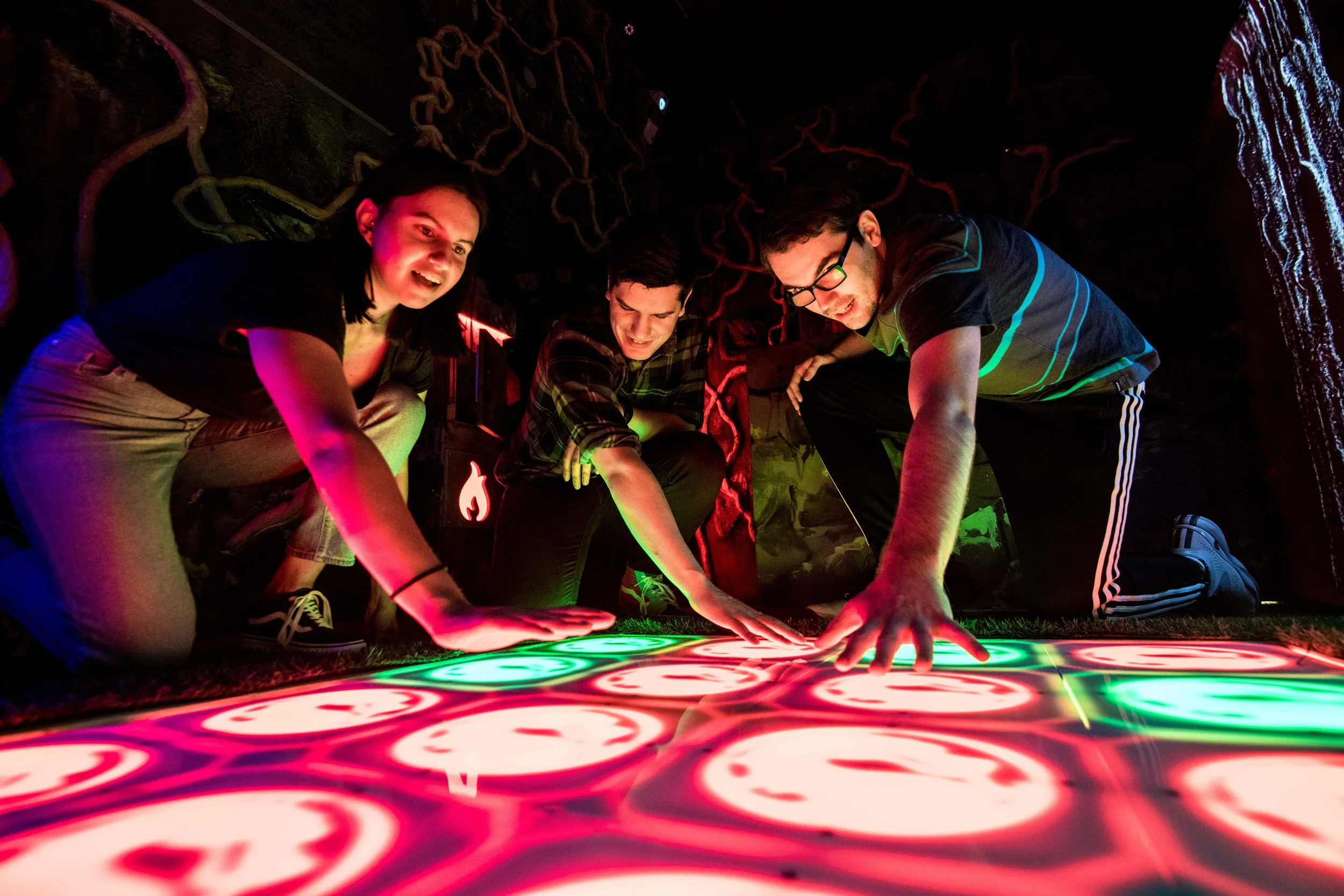 Three young adults engaging in an interactive light exhibit, with vibrant, swirling colored lights on the floor and a dark backdrop.