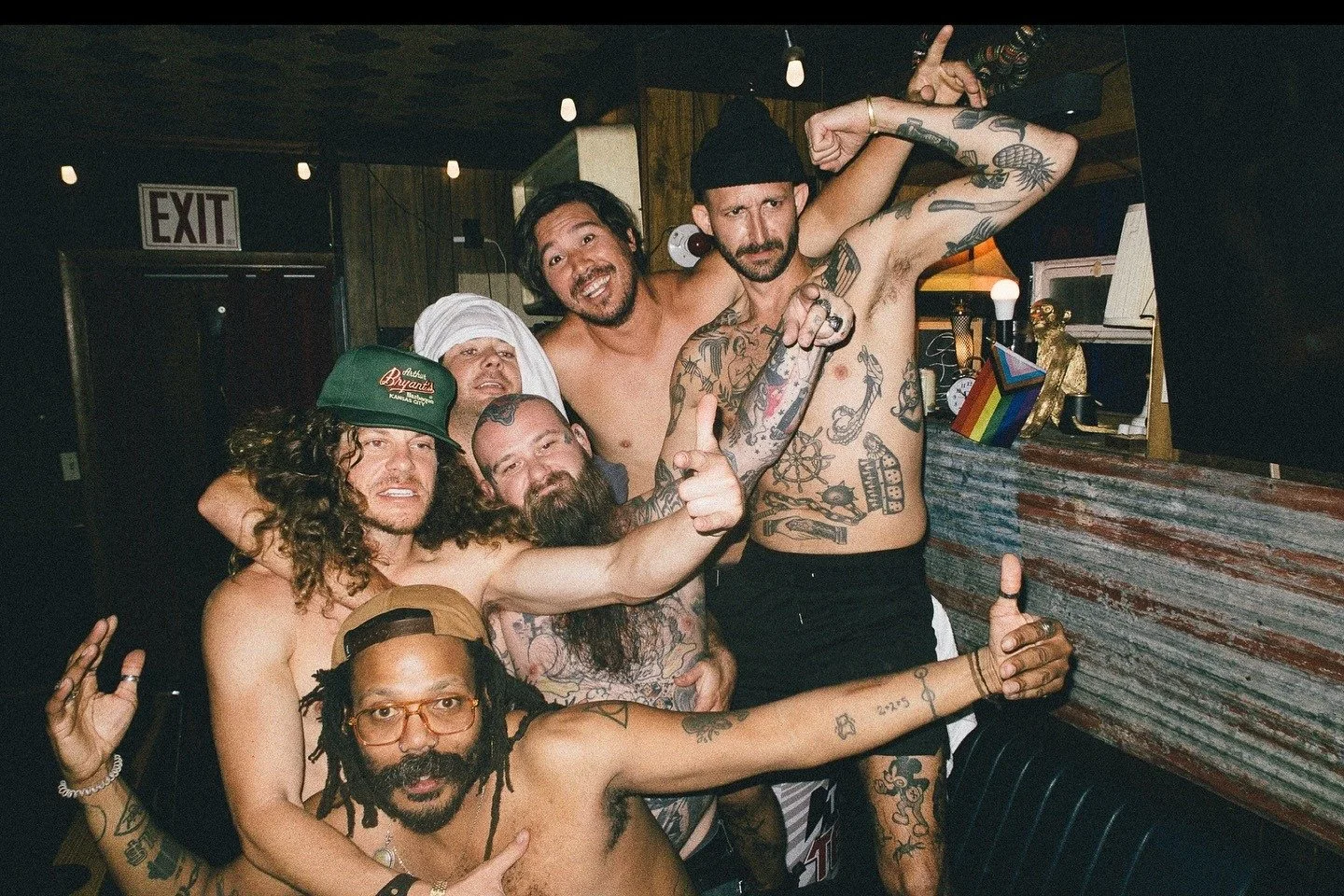 Group of shirtless men with tattoos celebrating, some making hand gestures, inside The Easy Inn bar Kansas City Kansas with wooden walls and a rainbow pride flag on the back counter. Pat Dougherty and Blake Anderson American actor and comedian