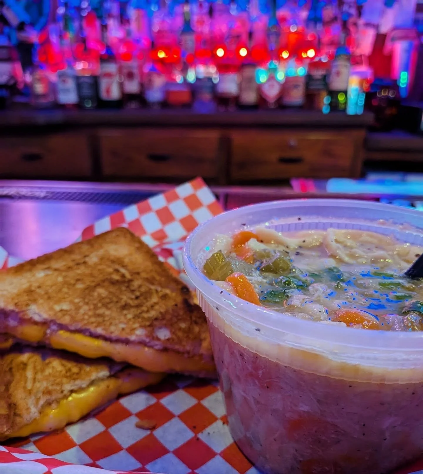 Close-up of a cup of soup, a grilled cheese sandwich, and a checkered paper in a cozy bar setting with colorful neon lights and bottles in the background at The Easy Inn Kansas Citys best kept secret underground dive bar serving great bar food