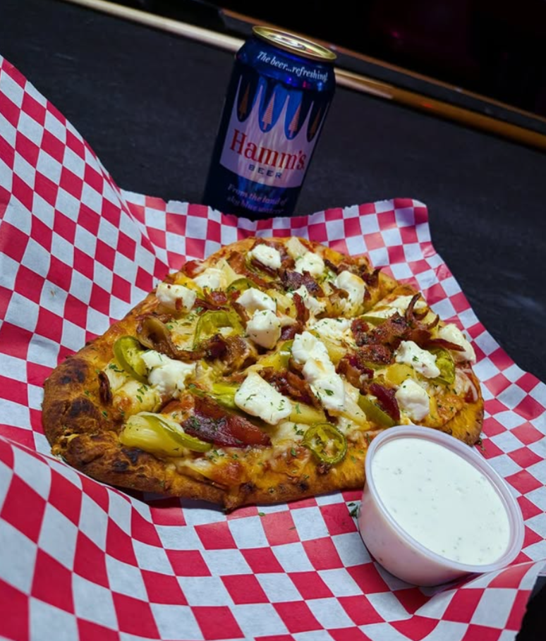 Slice of pizza with green peppers, bacon, tomatoes, and cheese, served on red and white checkered paper with a side of ranch dressing, a can of Hamm's beer in the background. The Easy Inn Kansas Citys best kept secret underground dive bar