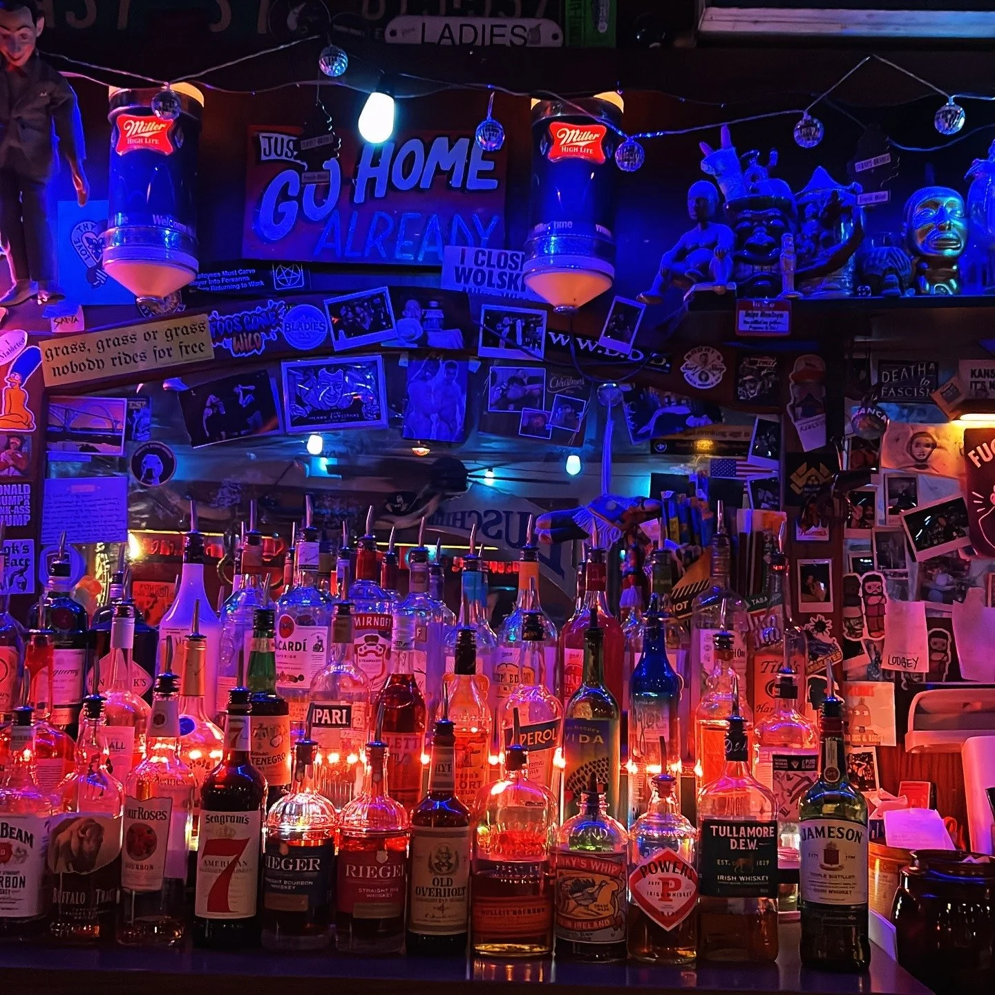 A bar scene with illuminated liquor bottles on the counter, illuminated by pink and blue neon lighting. The background features numerous posters, photos, and decorative items, including signs that say "Just Go Home Already," and various masks and figurines.