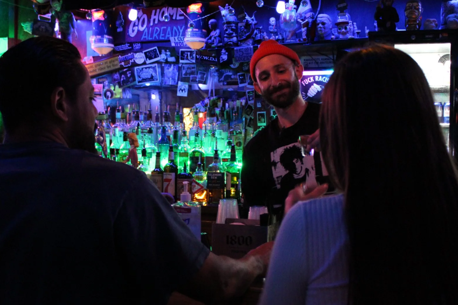 A dimly lit bar scene with three people conversing. The bartender, a man wearing a red beanie and a black shirt featuring a graphic, stands behind a counter filled with liquor bottles illuminated by green and purple neon lights. The bar's back wall is decorated with various posters, photographs, and figurines, including a 'Go Home Already' sign, and a neon sign reading 'F*** Racism' is visible.