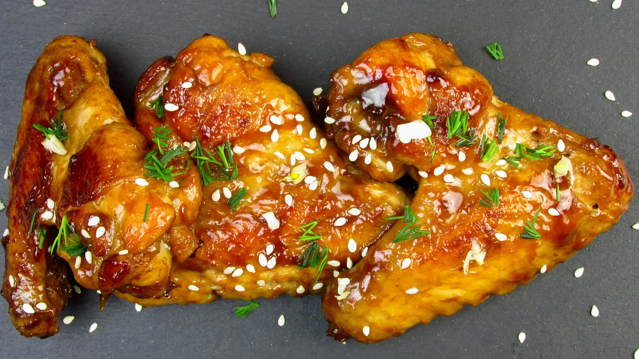 A cooked chicken drumstick and thigh glazed with sauce, garnished with sesame seeds and chopped green herbs on a dark surface.