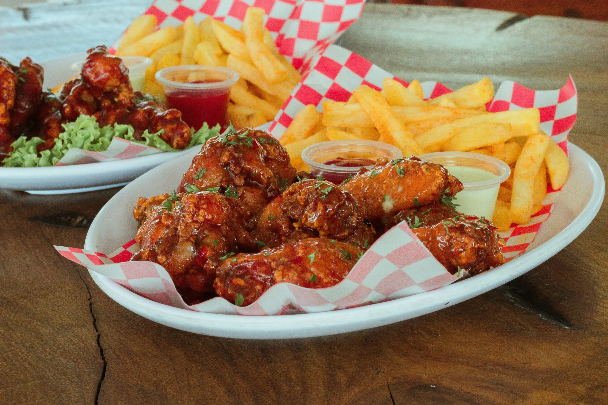 A basket of French fries, chicken wings with sauce and chopped parsley, and dipping sauces on a wooden table.
