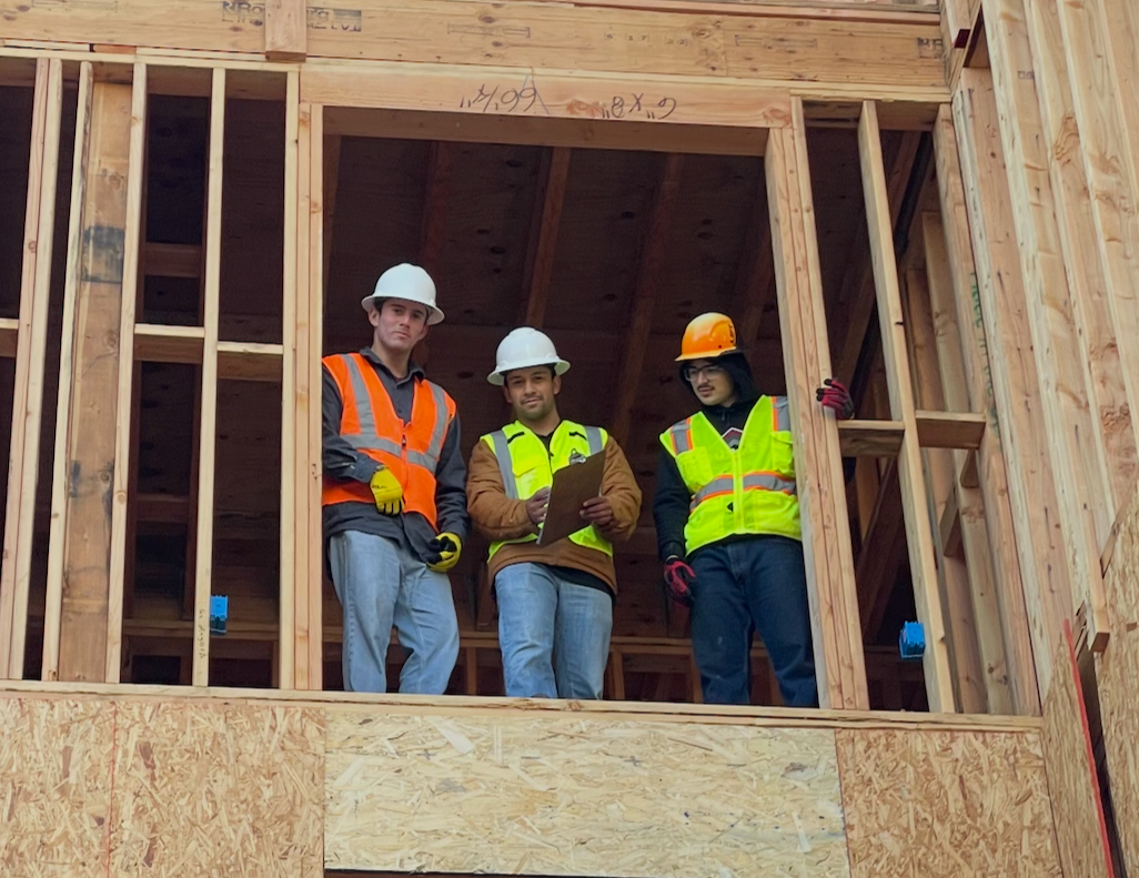 Three construction workers wearing safety helmets and vests standing on a wooden structure in a building under construction.