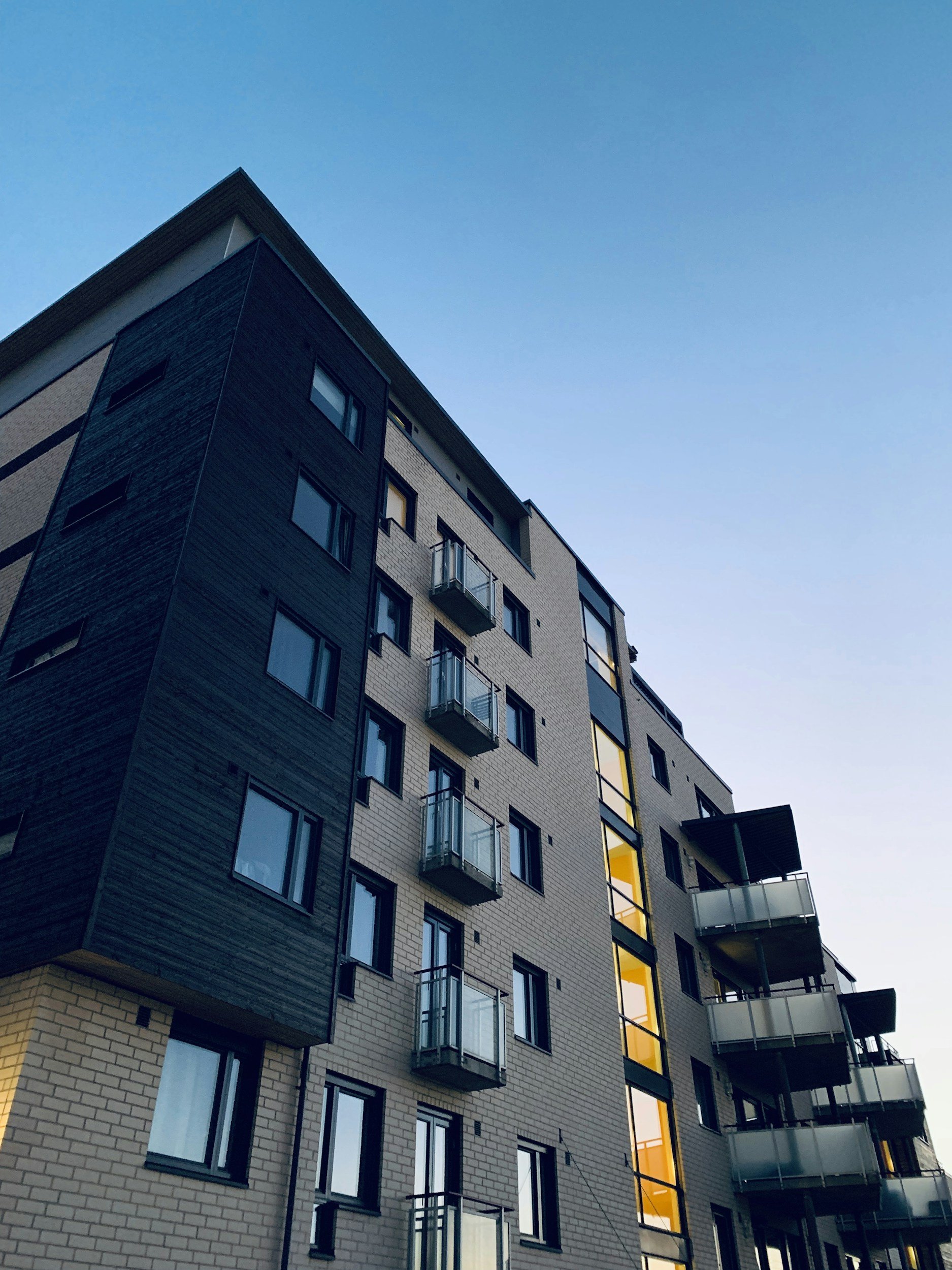 A multi-story apartment building with glass balcony railings, black, white, and beige brick facade, and lit interior windows against a clear blue sky.