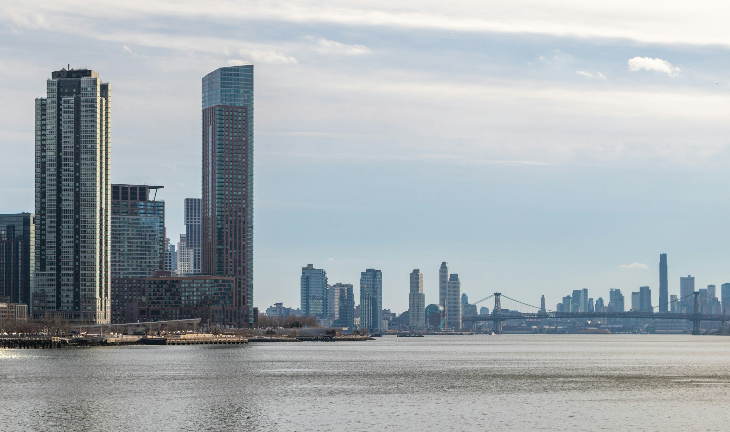 City skyline with tall modern skyscrapers, a bridge, and water in the foreground under a partly cloudy sky.