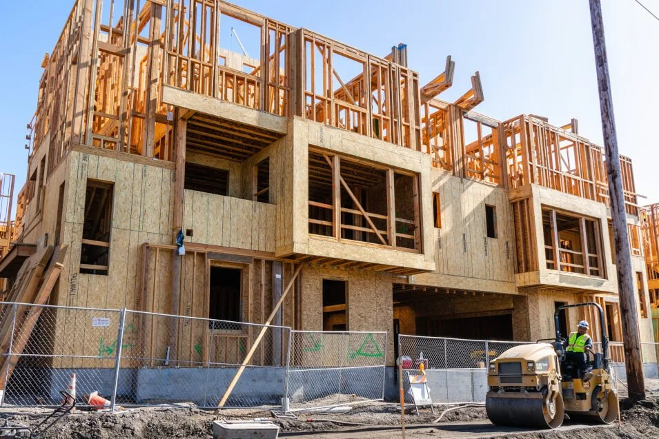 Construction site of a multi-story building with wooden framing and concrete walls, surrounded by a safety fence. A worker in a safety vest and helmet operates a road roller in the foreground, and construction materials and equipment are scattered around.