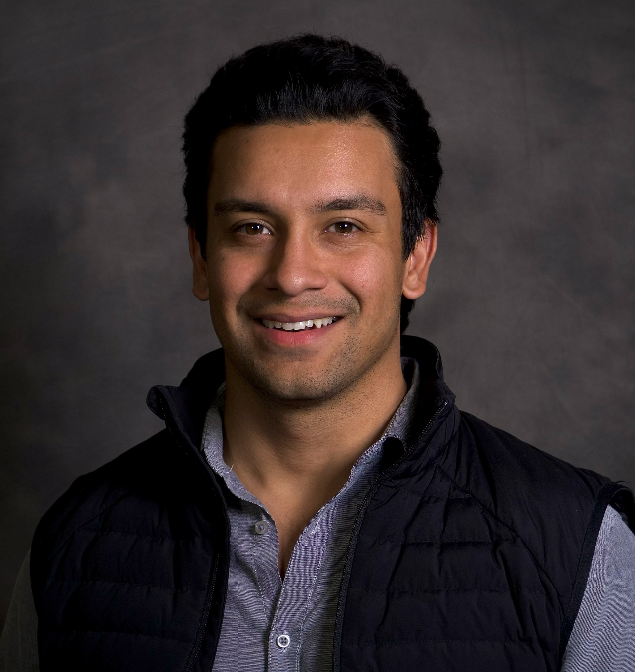 Headshot of a smiling man with dark hair, wearing a gray button-up shirt and black vest against a dark background.