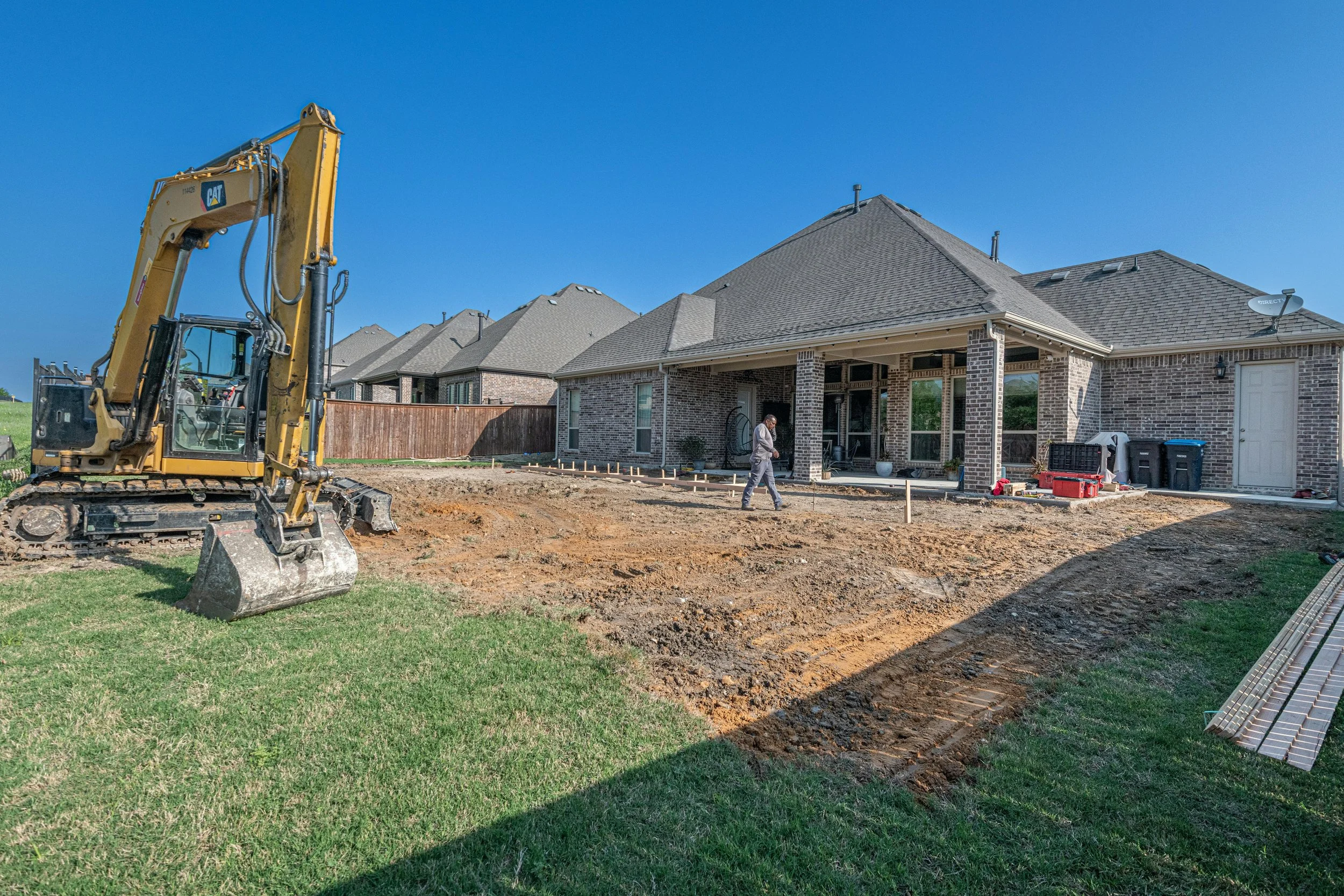 A new backyard with freshly tilled soil and a row of houses in the background. There is construction equipment and tools, and a person working in the yard.