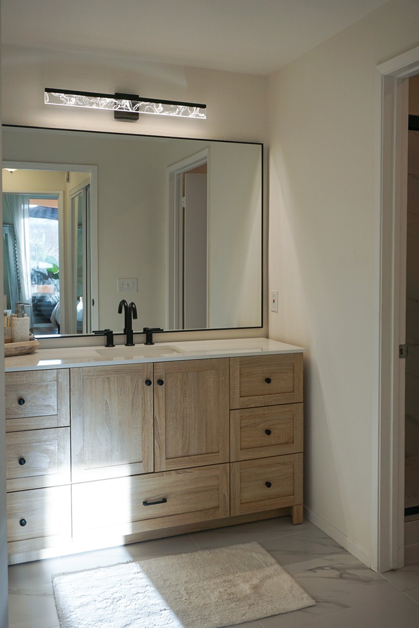 Bathroom with wooden vanity, white countertop, black faucet, large mirror, wall-mounted light, light-colored walls, and a cream rug on gray marble floor.