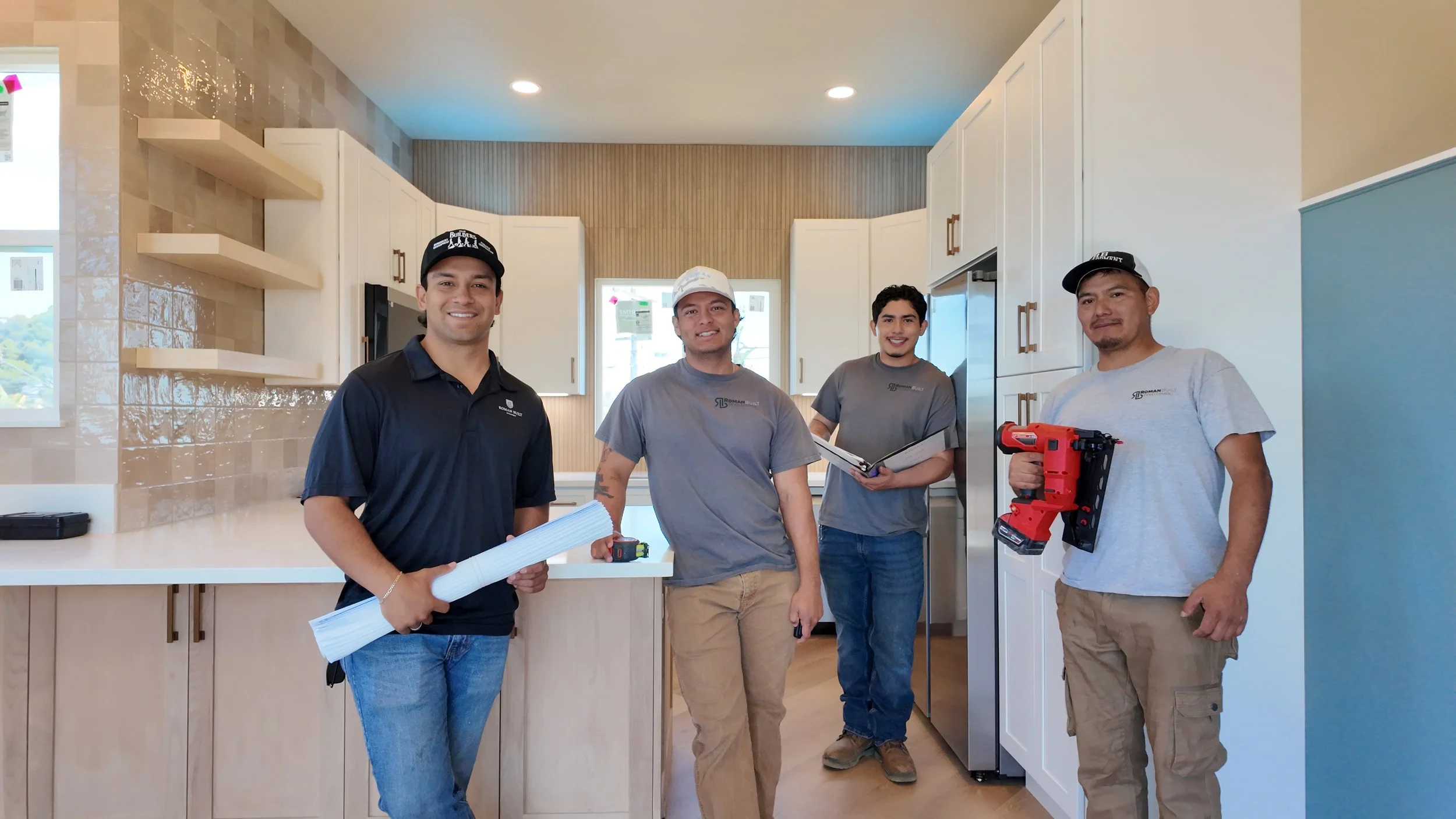 Four men standing in a kitchen, holding tools and documents, smiling at the camera.