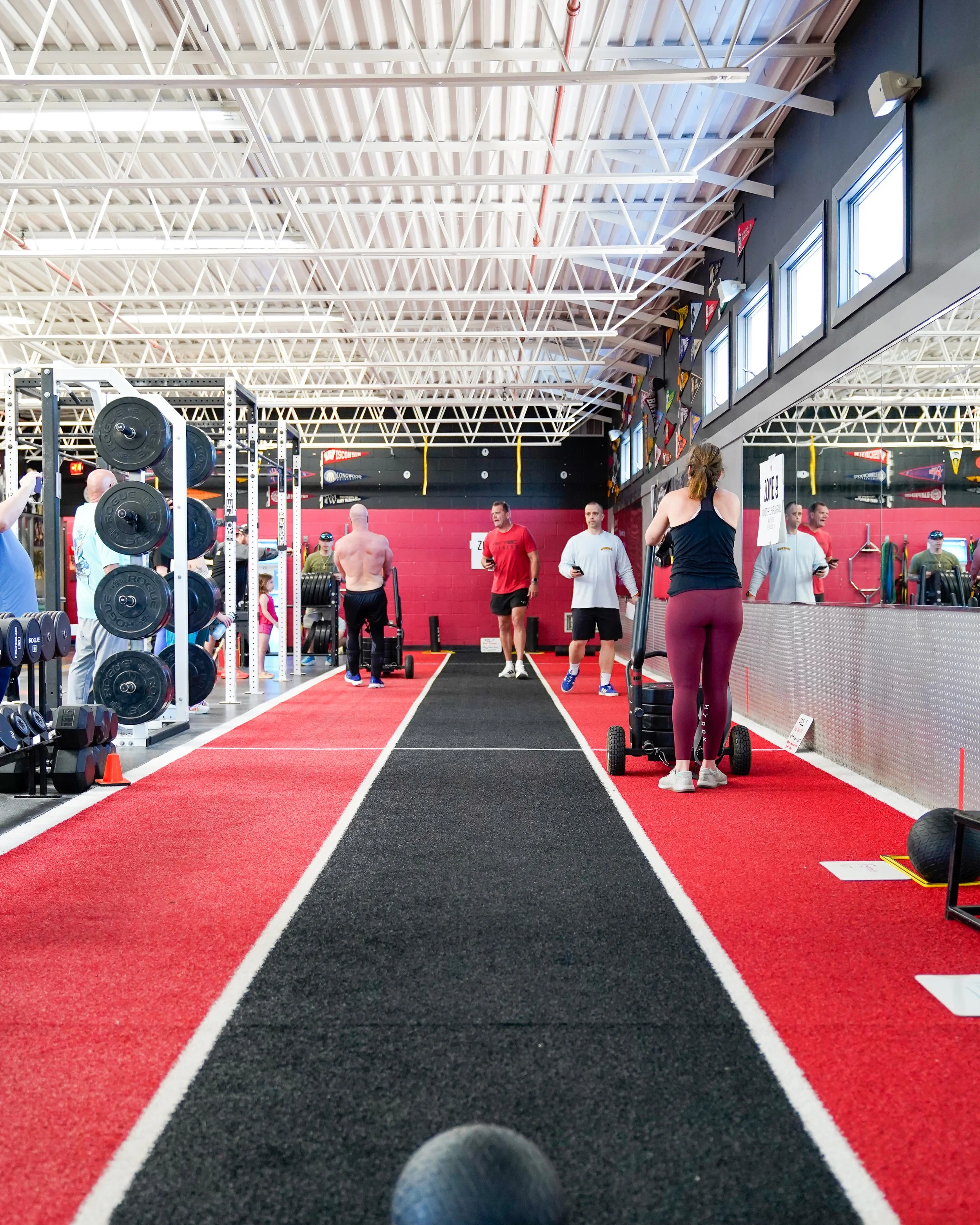 People working out in a gym with black and red flooring, weight racks, and fitness equipment, viewed from a perspective of a person holding a macebell.