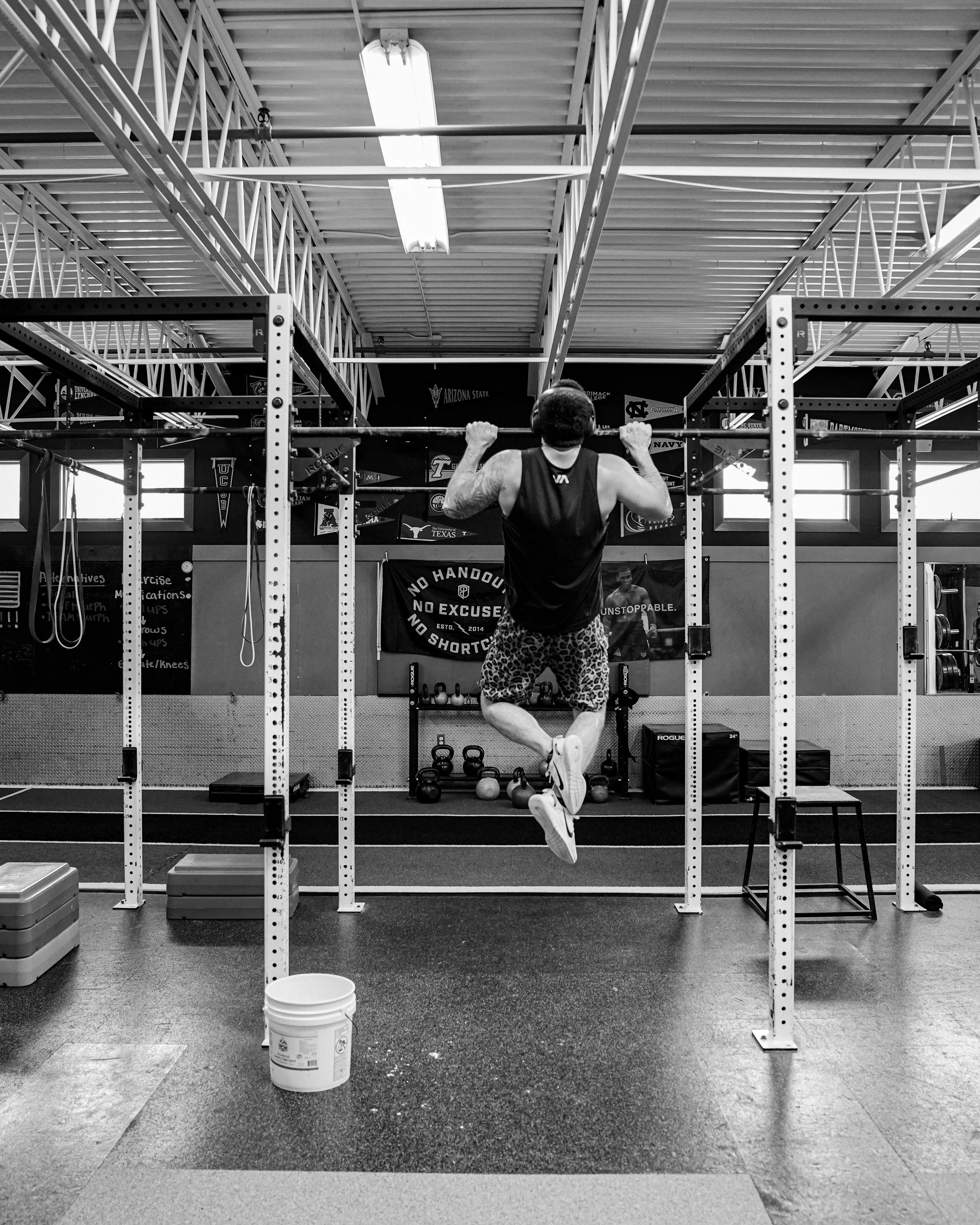 A man doing pull-ups in a gym, with weights on the floor and gym posters on the wall.