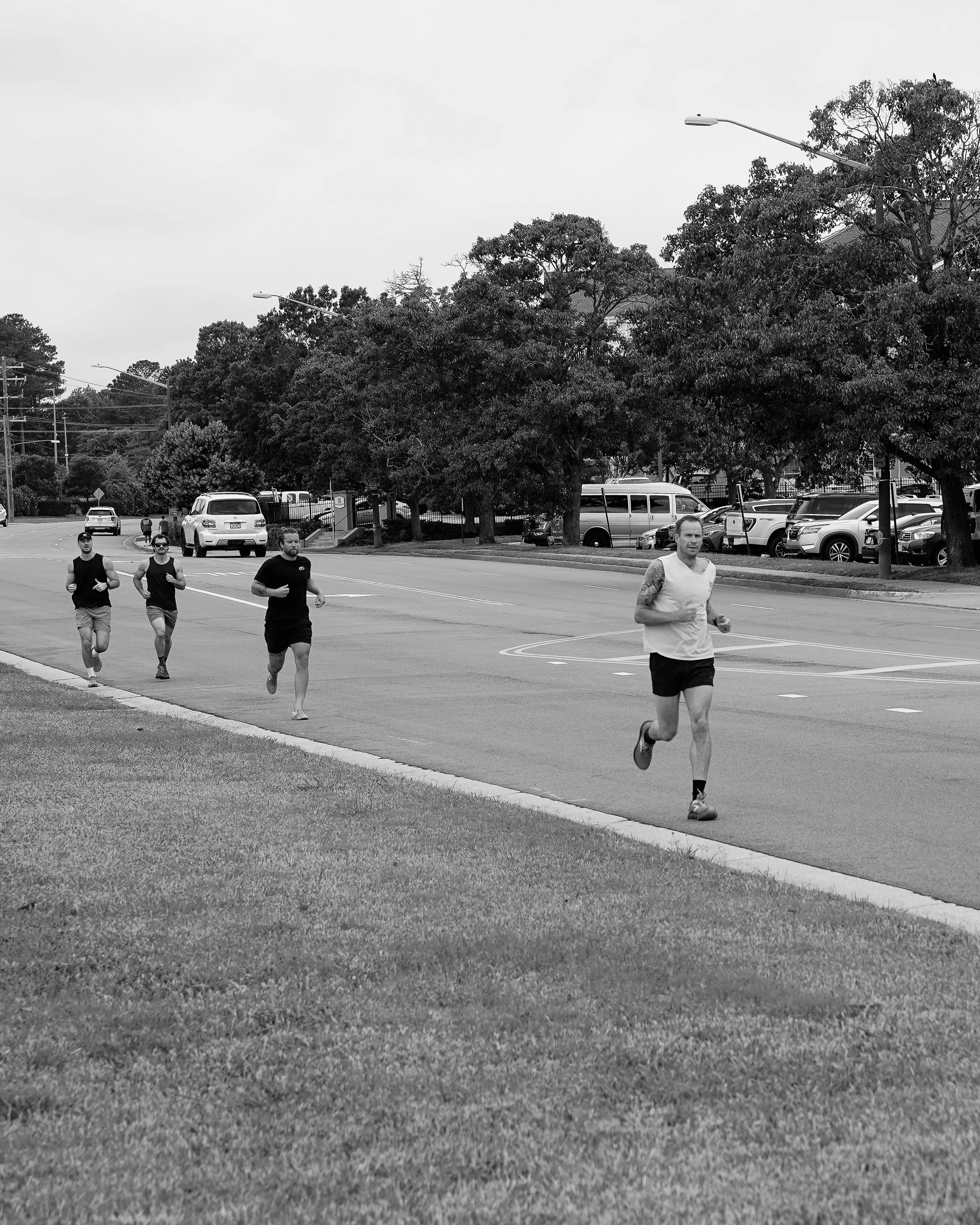 Four people jogging along a street with parked cars, trees, and streetlights in the background, in a black and white image.