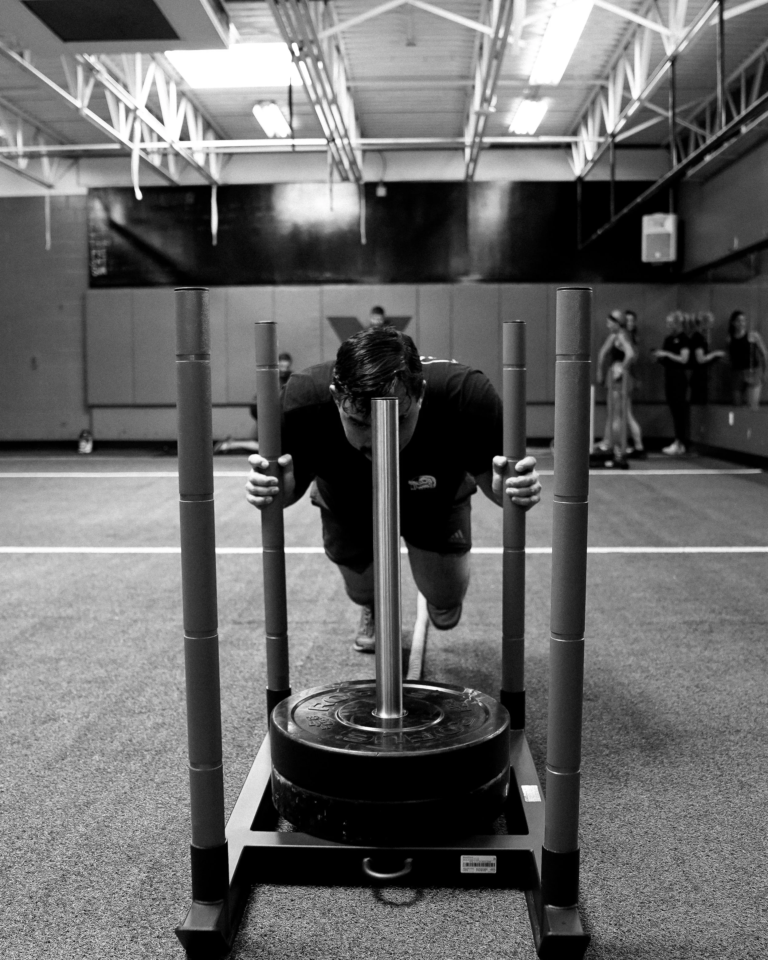 A person in black workout clothing is in a gym, performing a push-up exercise on a black rubber mat with their hands on a metal barbell. The gym has a high ceiling with fluorescent lights, and several other people are visible in the background.