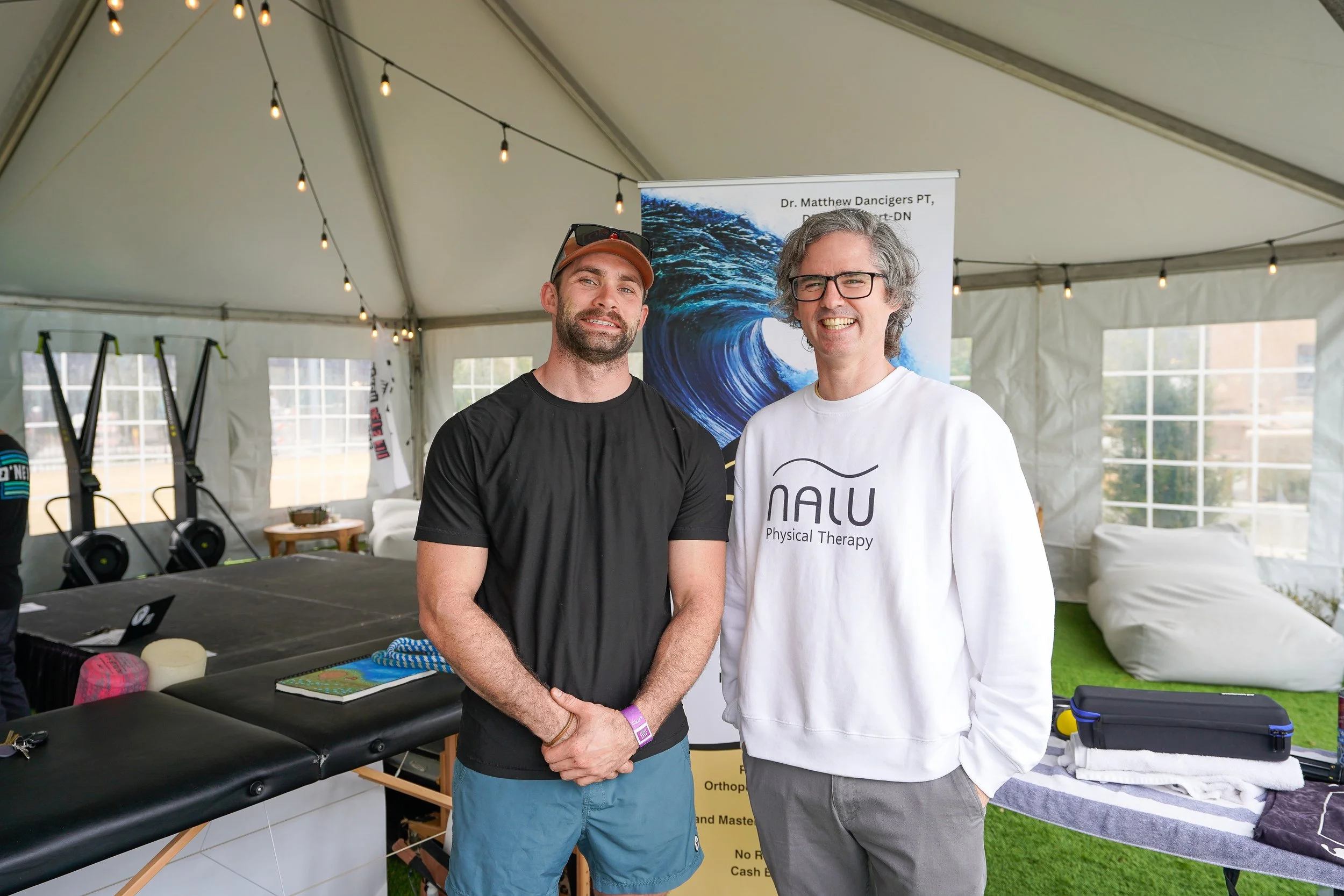 Two men standing inside a tent at an event, smiling at the camera. One man is wearing a black t-shirt and a baseball cap, the other a white shirt with text. There are lighting strings overhead, and a table with various items behind them, including a 