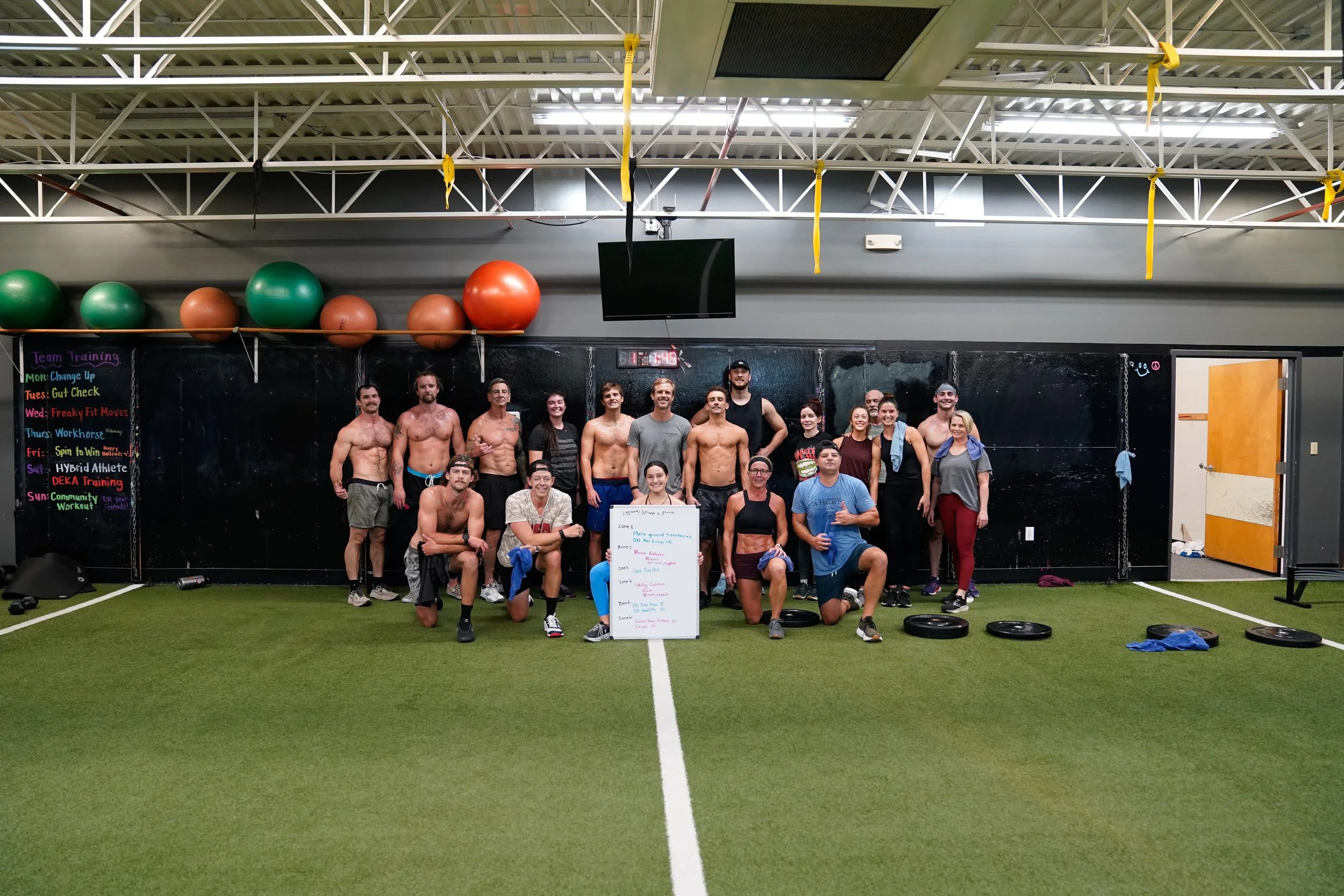 A group of fitness trainers and participants posing for a photo in a gym with green turf flooring, large exercise balls on a shelf, a digital clock, and a whiteboard with workout zones written on it.