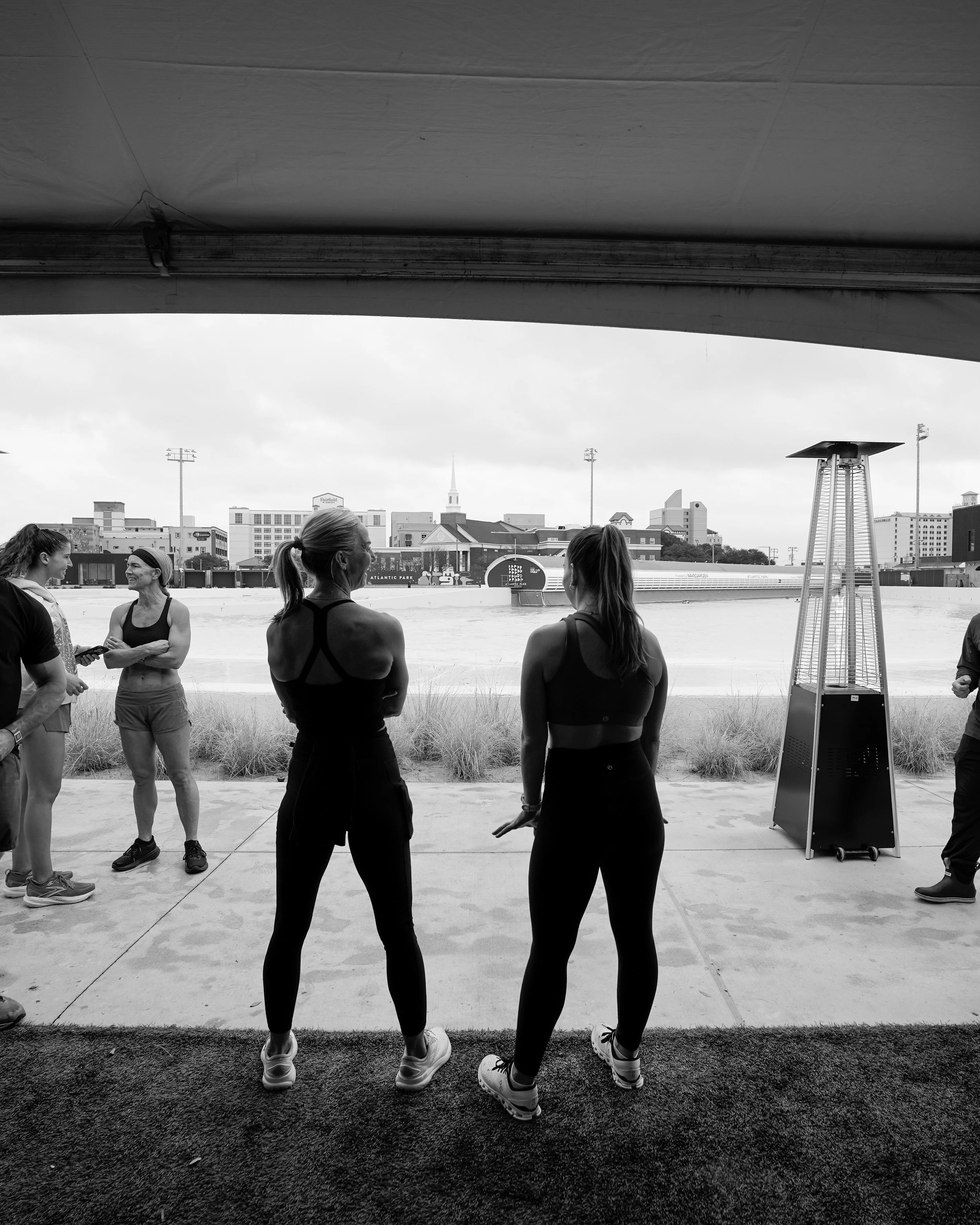 Four women in workout clothes are standing outdoors on a cloudy day, facing each other and talking, with a city skyline in the background. A tall patio heater is to the right.