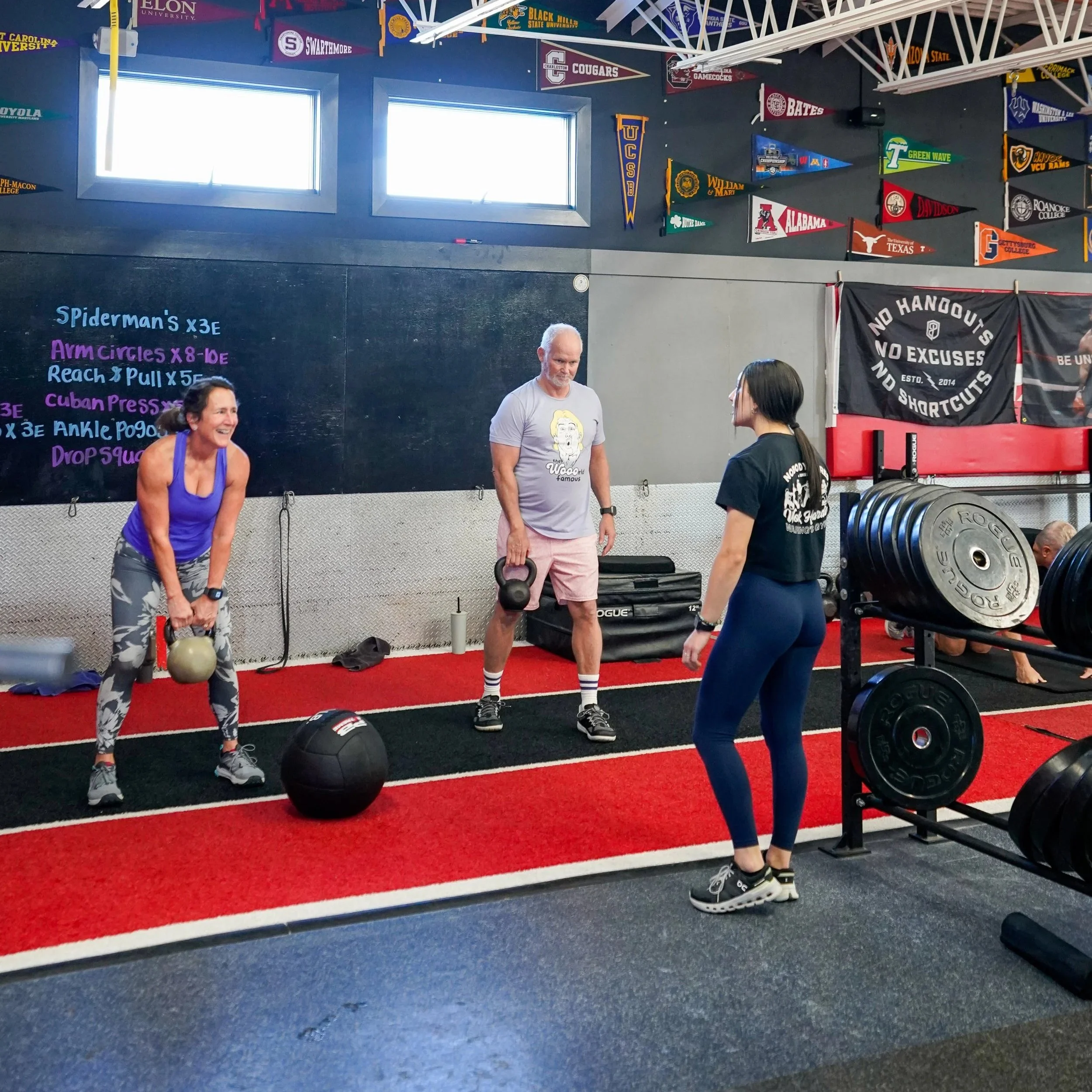 Group of people working out in a gym with weights, banners hanging on the wall, and a brightly lit window.