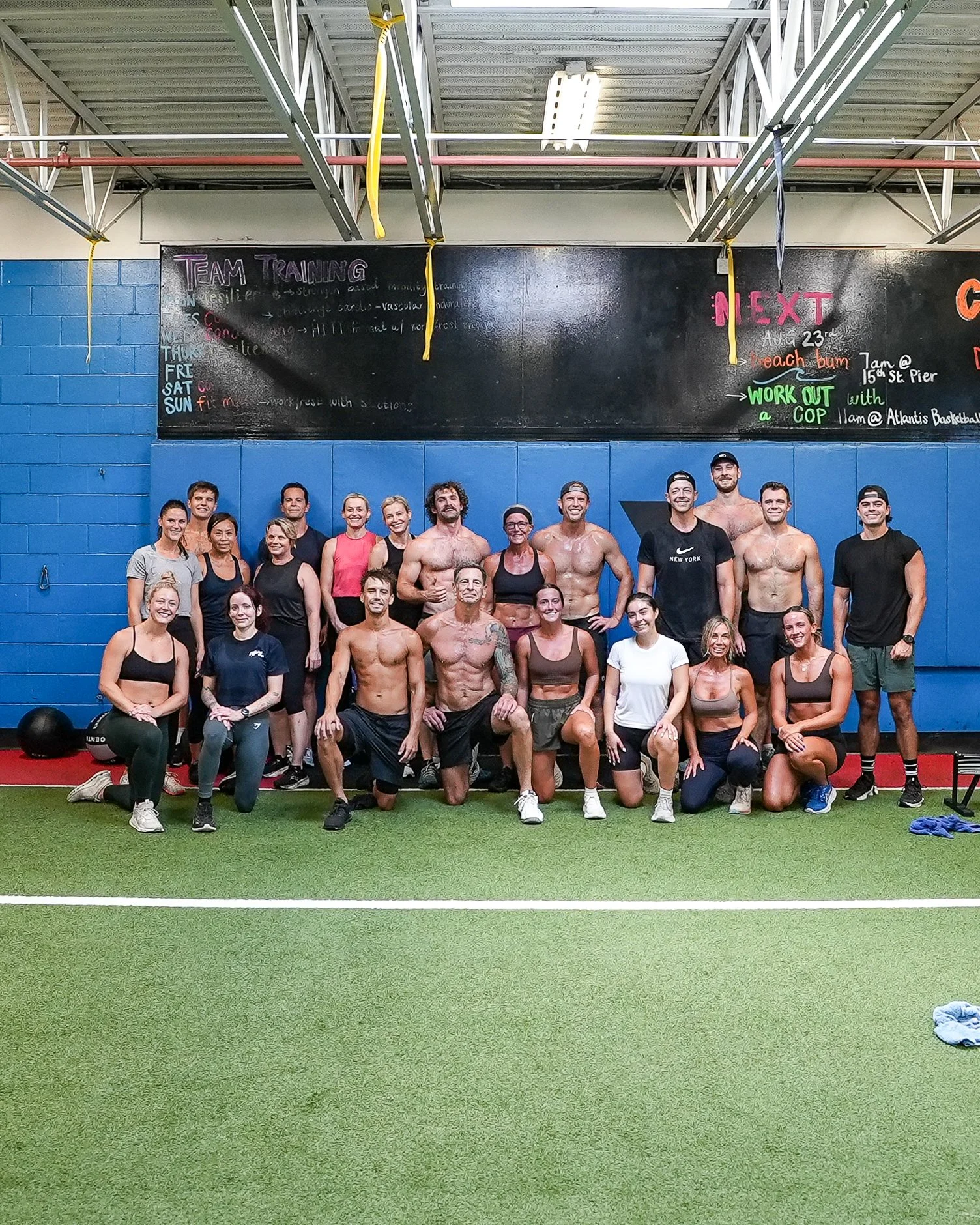 A large group of people, both men and women, posing for a photo inside a gym with a blue wall and blackboard above. Some are standing, some kneeling or sitting. The group appears to be in a fitness or workout class.
