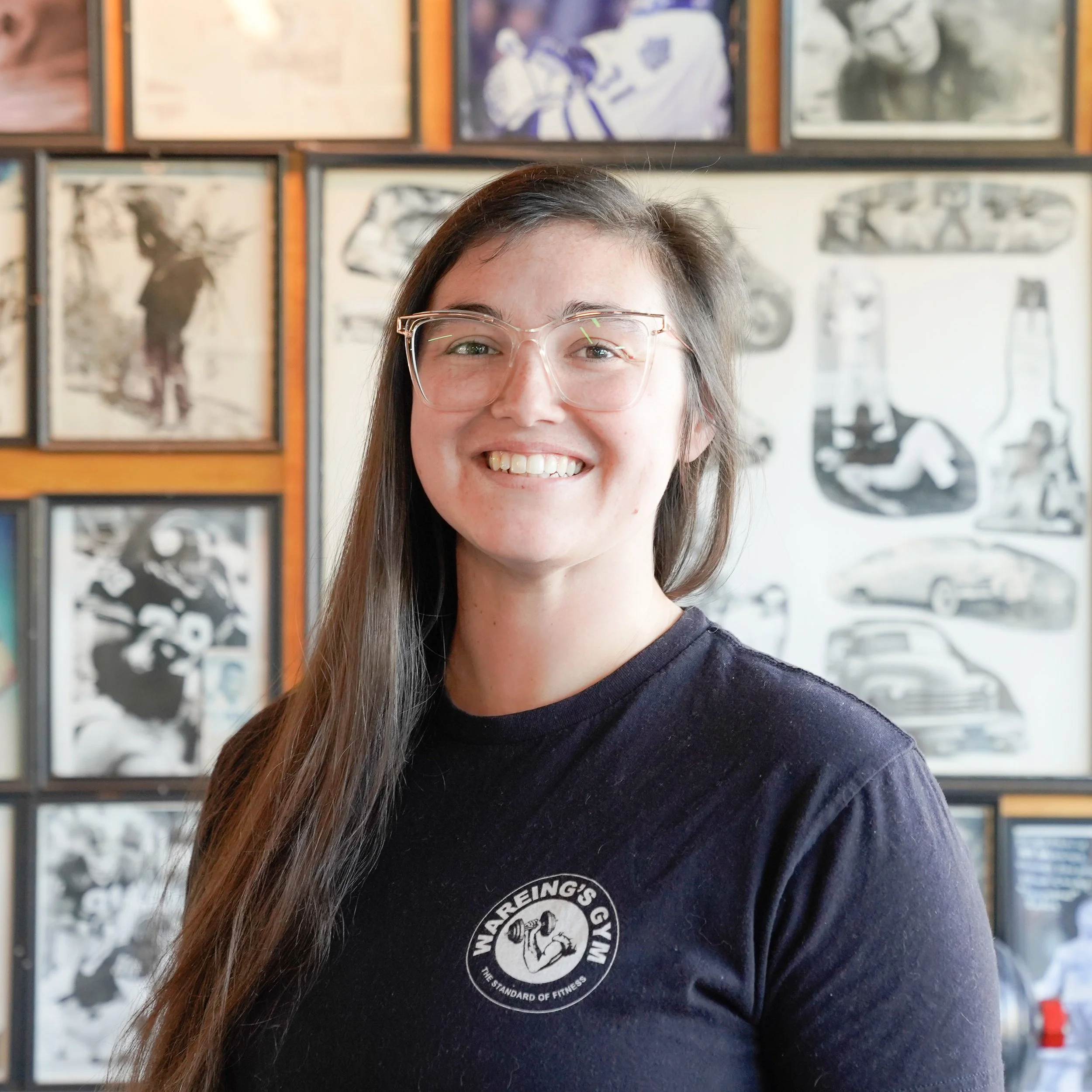 A woman with long brown hair and glasses smiling in front of a wall of black and white framed artwork, wearing a black shirt with a circular logo that reads "WARRING'S GYM, THE STANDARD OF FITNESS."