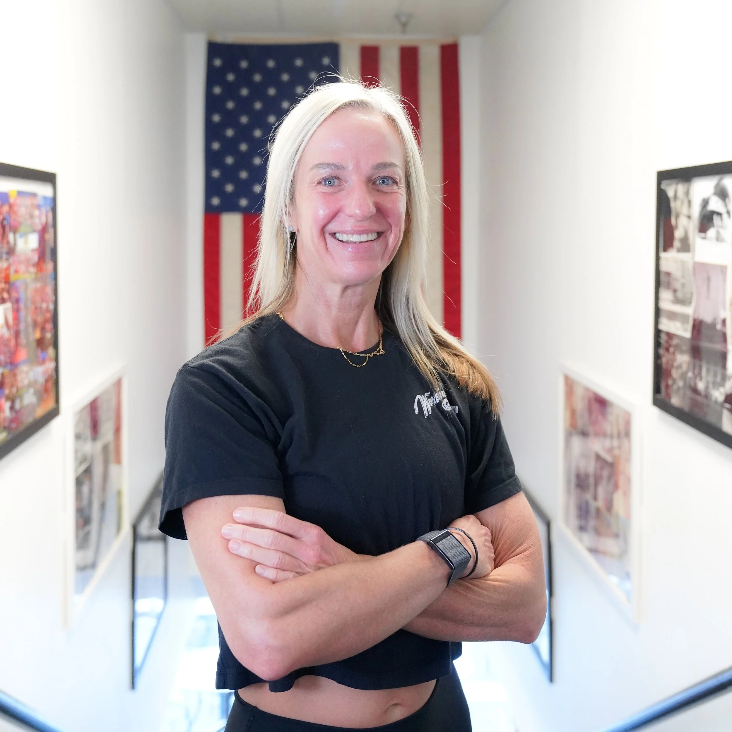 A woman with long blonde hair smiling, crossing her arms, standing in front of an American flag hanging on the wall, with pictures on the side walls.