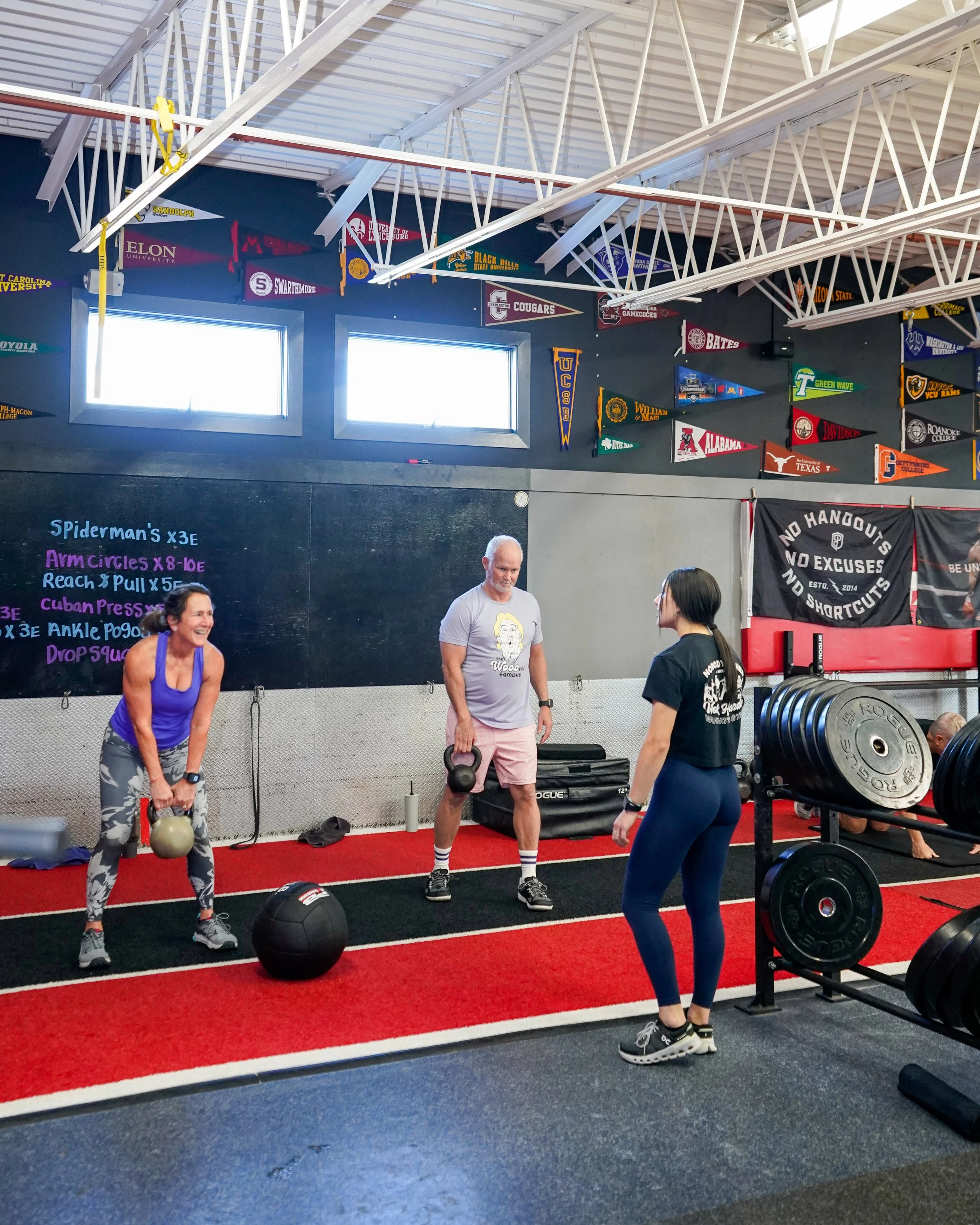Indoor gym with people working out. Three people near weights and exercise equipment. One woman with long hair in a blue tank top and camo leggings, another man with a kettlebell and a woman with a black t-shirt. Gym has banners hanging on the wall, 