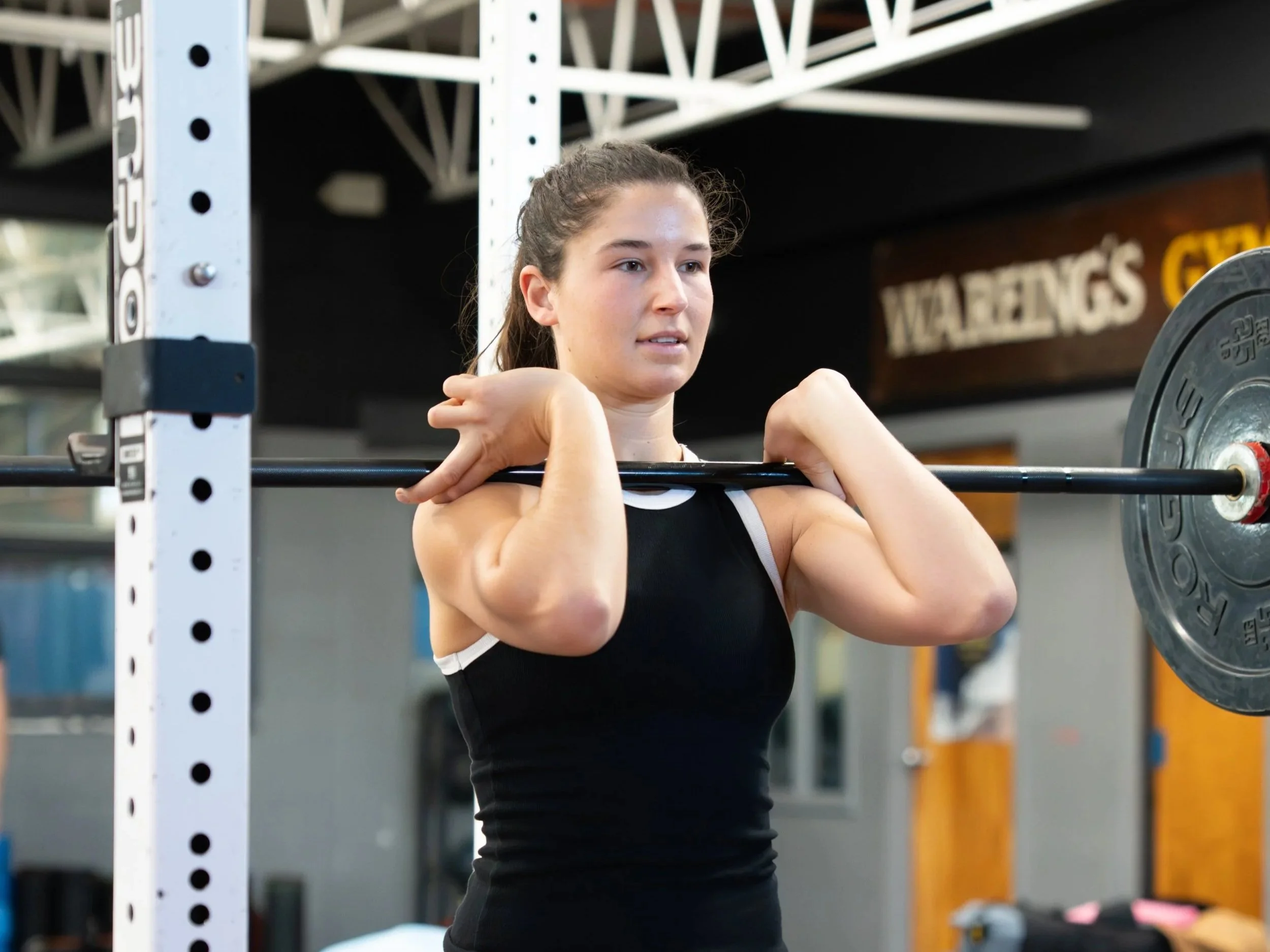 A young woman with brown hair tied back in a ponytail, wearing a black athletic singlet, is lifting a barbell in a gym. The gym has a structure with white metal beams and weights in the background.