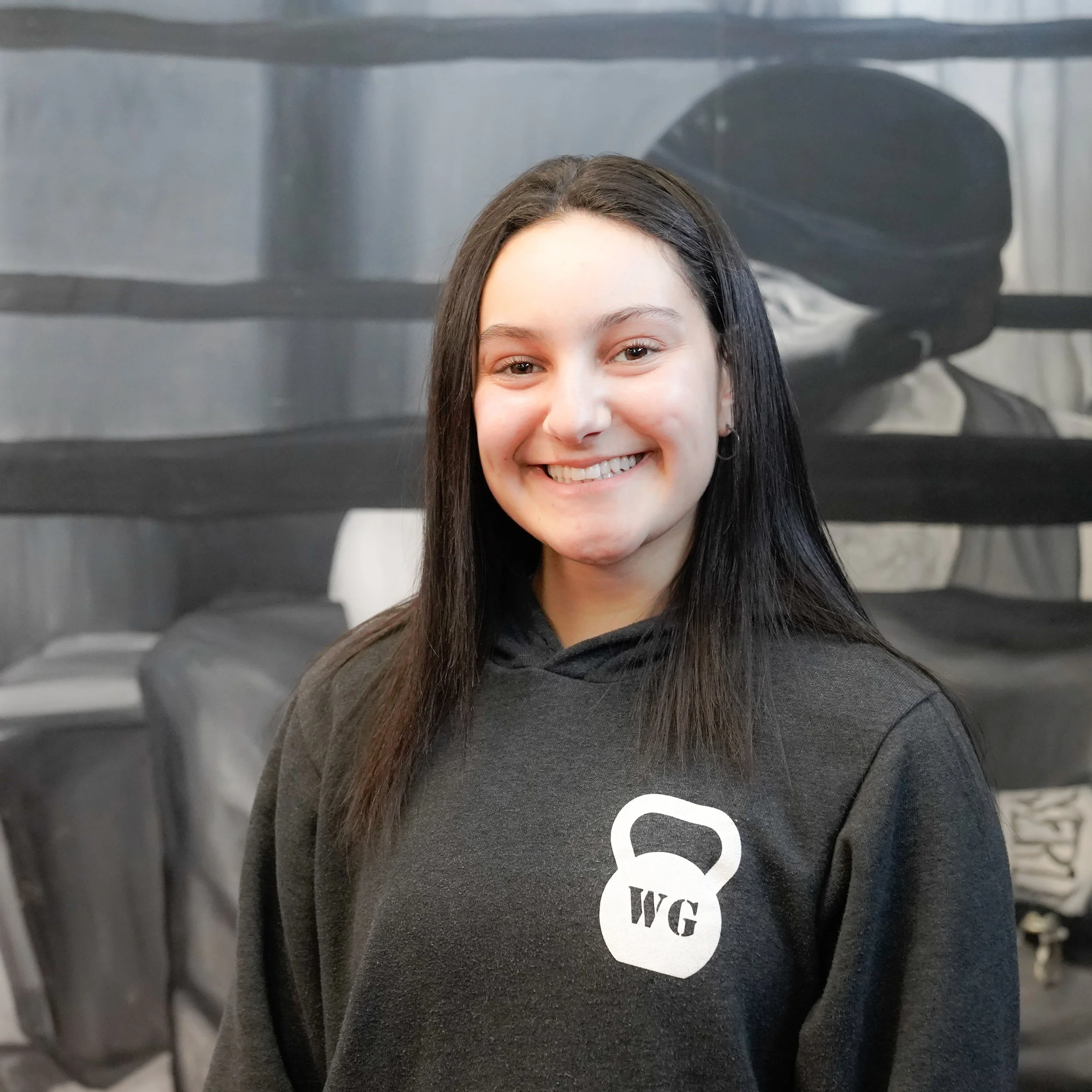 Close-up of a young woman with long dark hair and a bright smile, wearing a black hoodie with a white graphic logo, standing in front of a wall of black t-shirts displayed on racks.