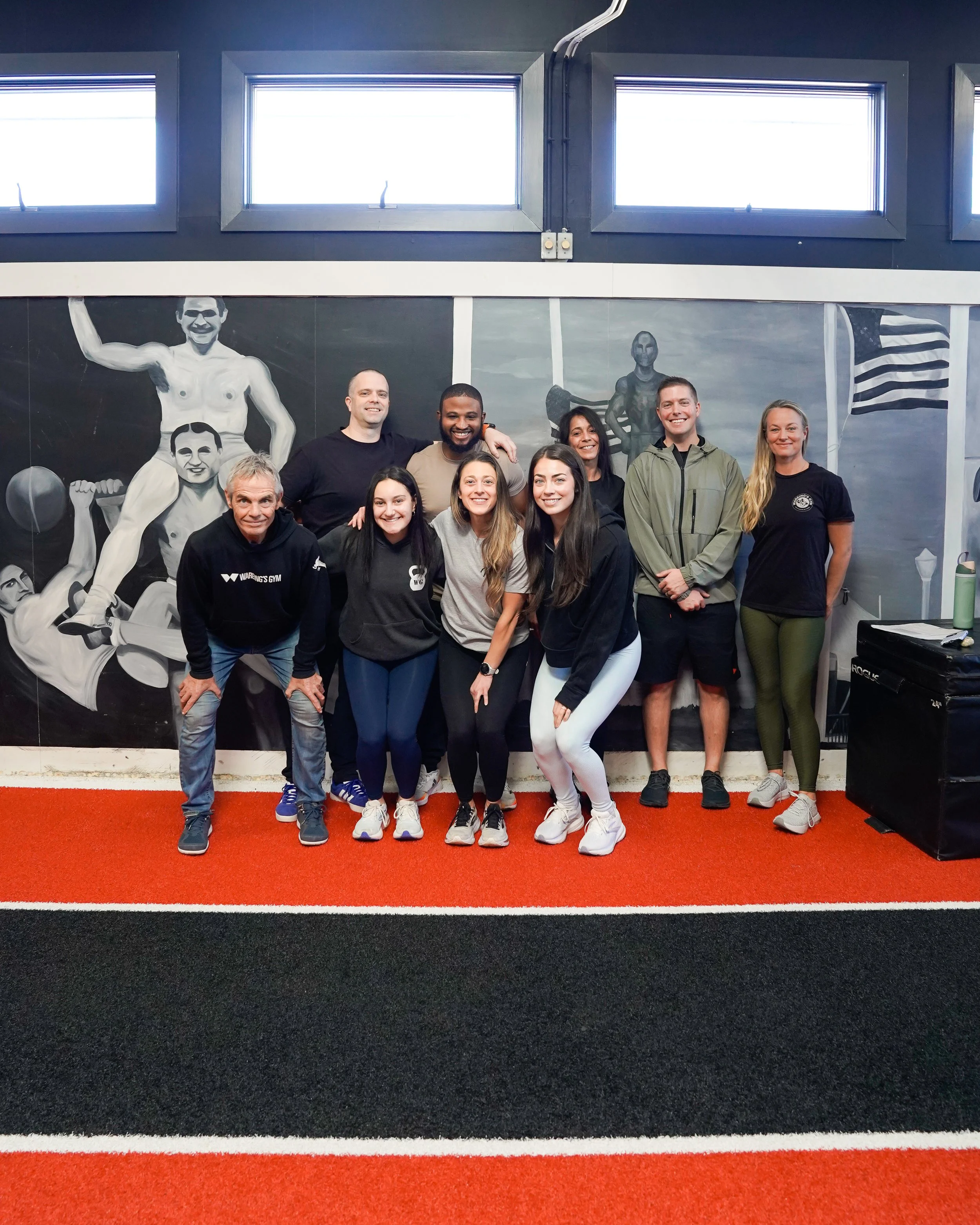 Group of nine people posing in front of a black and white mural of fitness figures inside a gym. The floor has a red and black color scheme, and there are three windows near the ceiling.