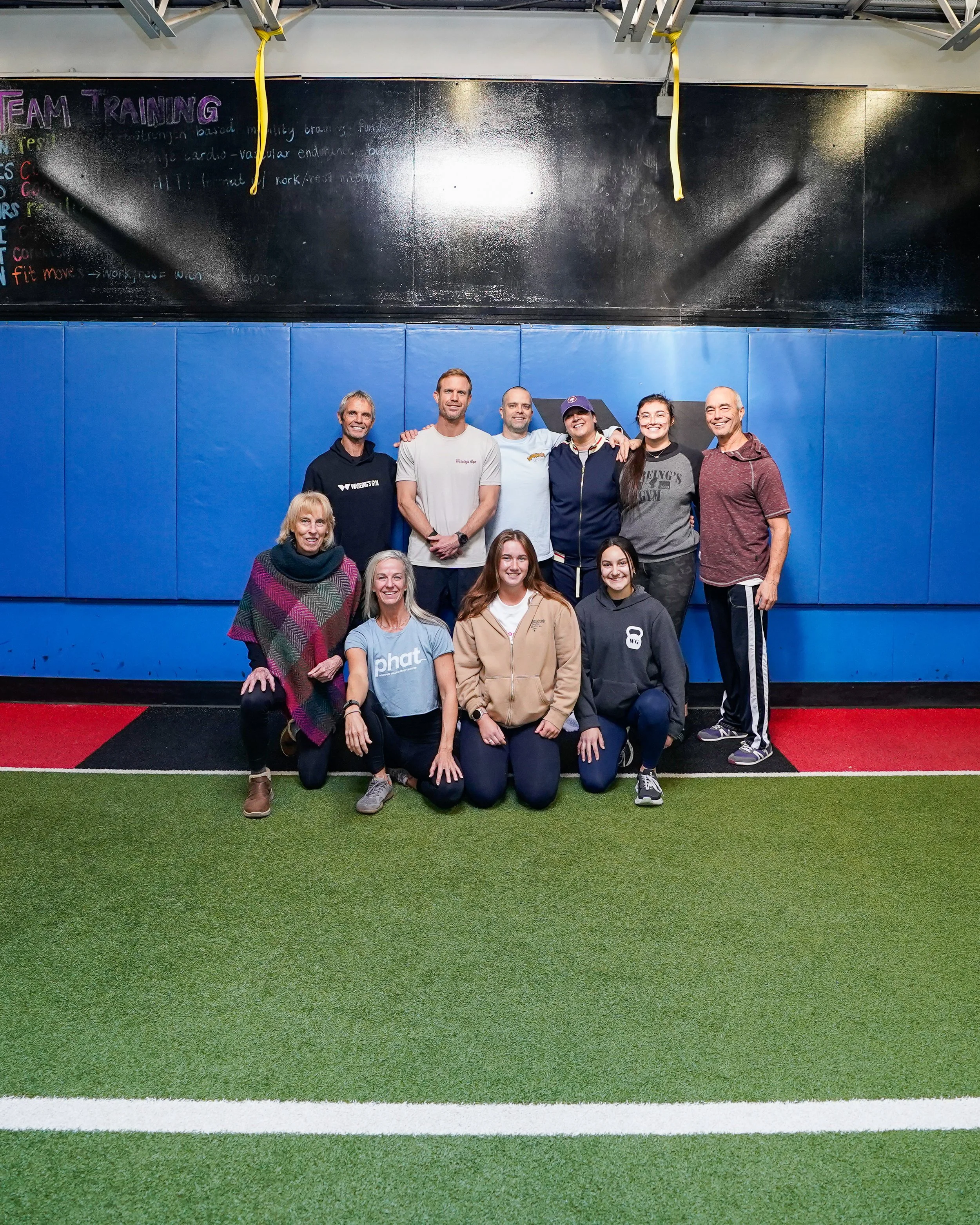 A group of nine people, including adults and children, standing together on a sports field with artificial turf, smiling for a photo, in front of padded blue walls and a blackboard with writing about team training.