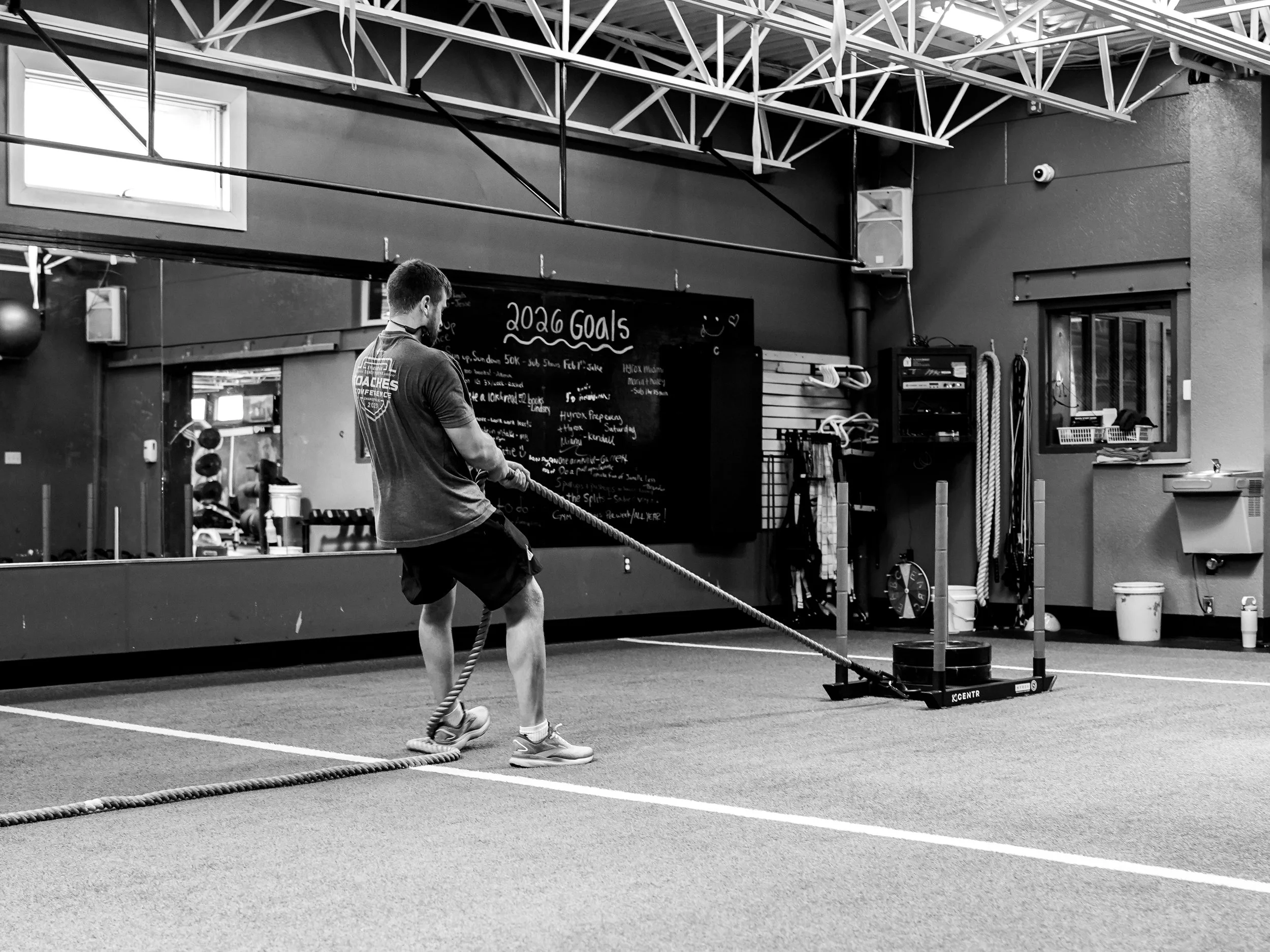 A man in athletic attire pulls on a battle rope in a gym with black walls, a blackboard with '2026 Goals' written on it, and fitness equipment like kettlebells and resistance bands.