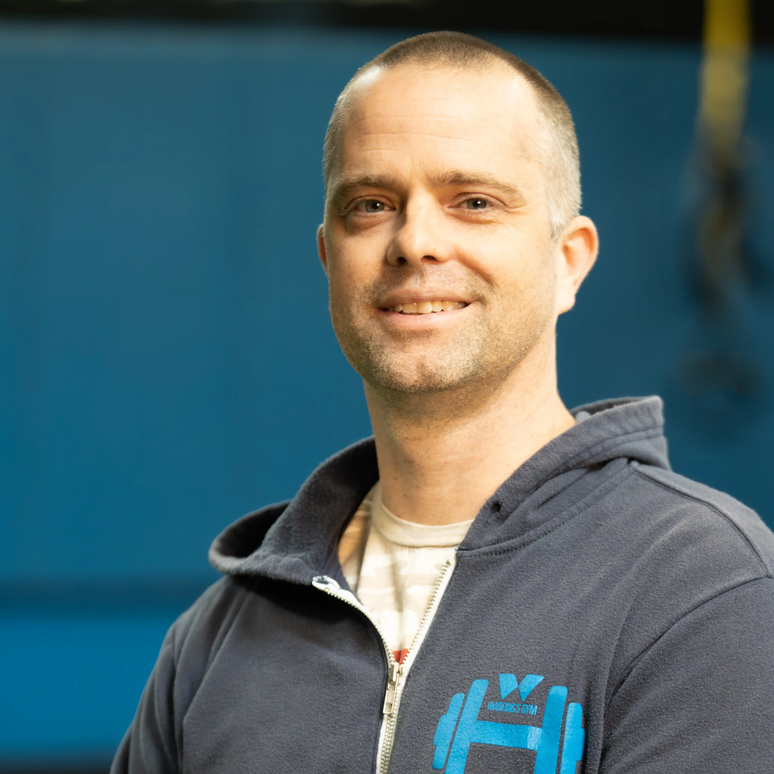 Close-up of a smiling man with light skin and short hair, wearing a dark hoodie over a white t-shirt, standing against a blue background.