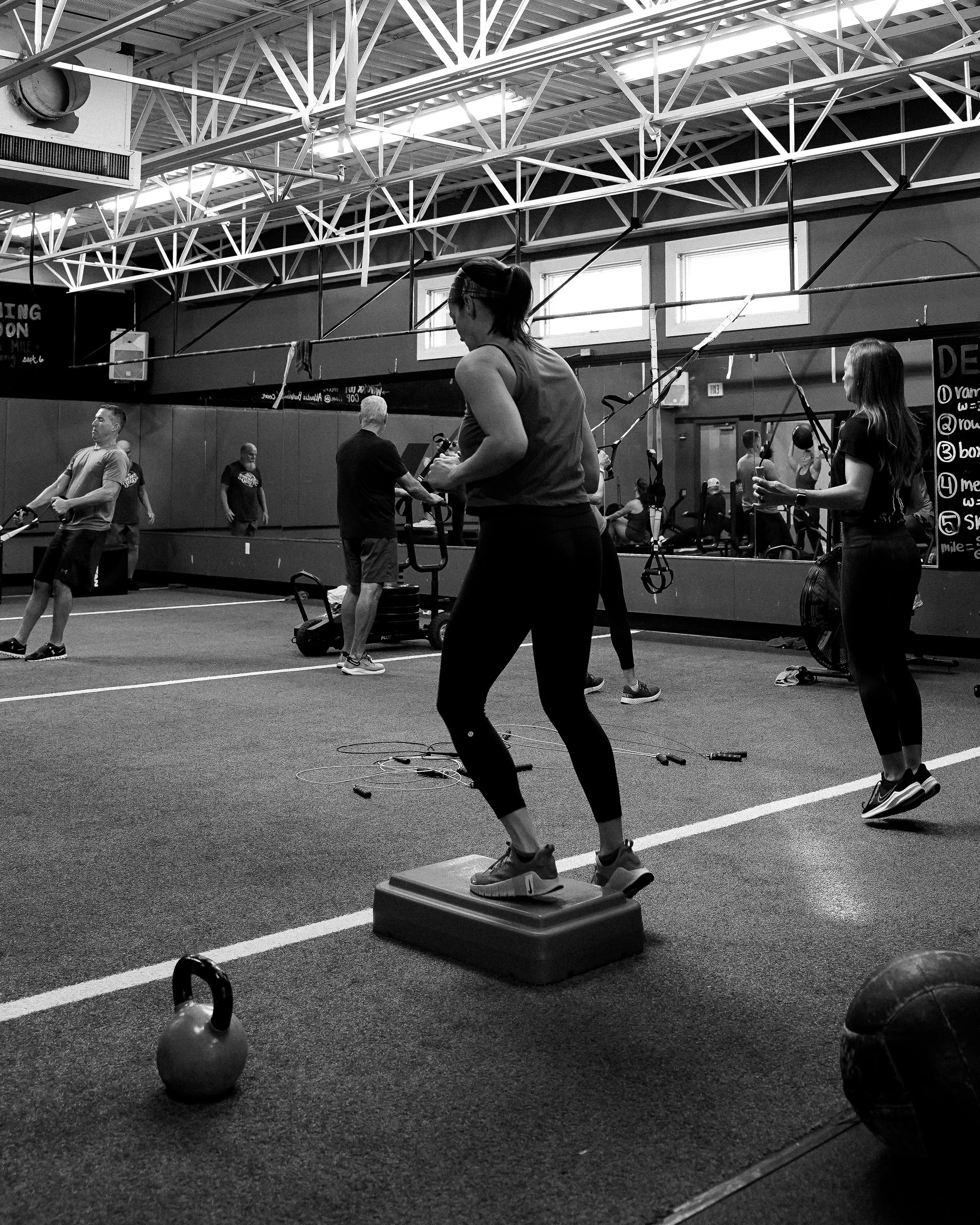 A woman exercising with resistance bands tied to the ceiling in a gym, standing on a step platform. Other people are working out in the background, with kettlebells and gym equipment visible.
