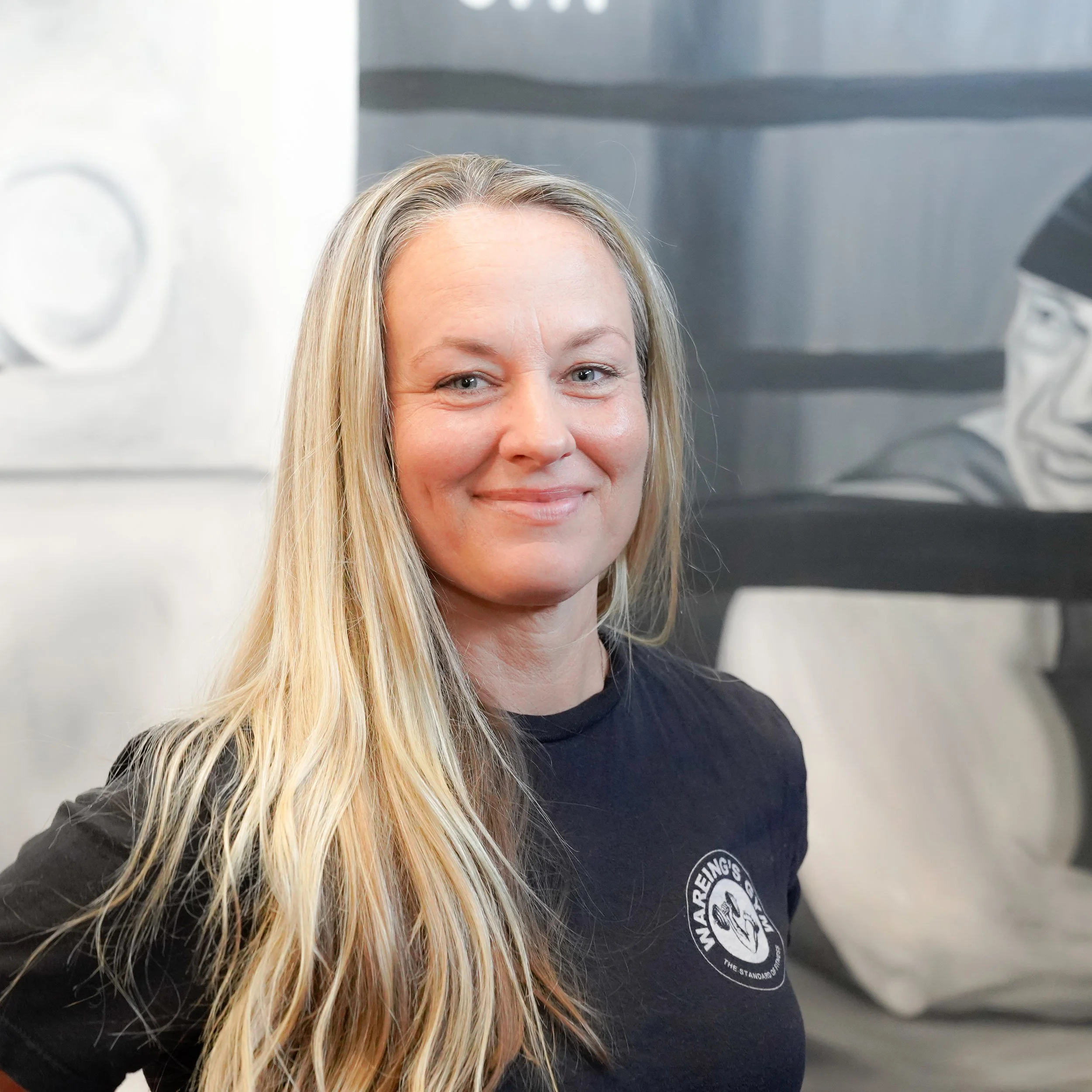 Close-up of a woman with long blonde hair smiling, wearing a black shirt with a circular patch on it.