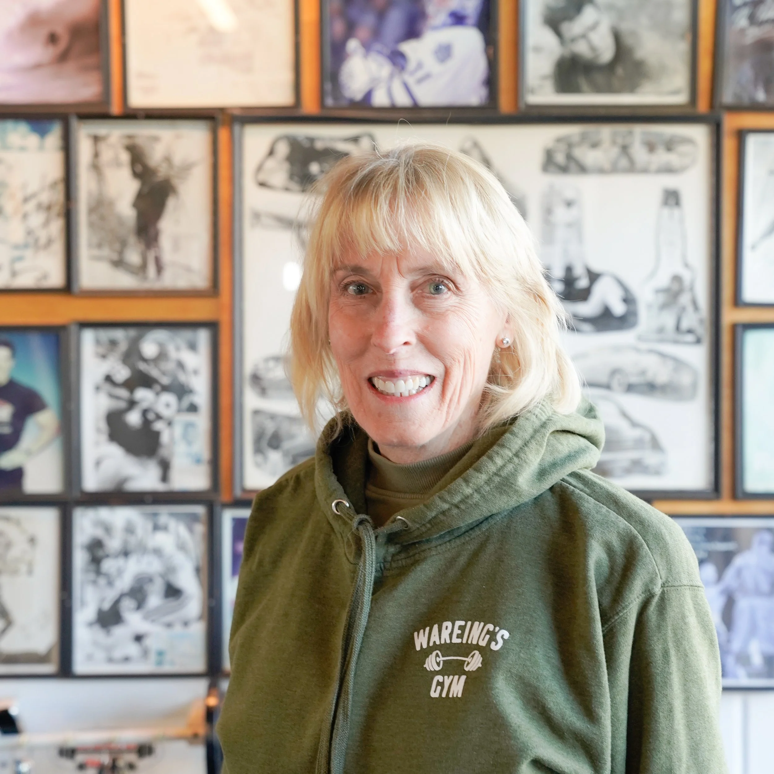 Close-up of a smiling woman with blonde hair, wearing a green hoodie, in front of a wall with black-and-white photographs.